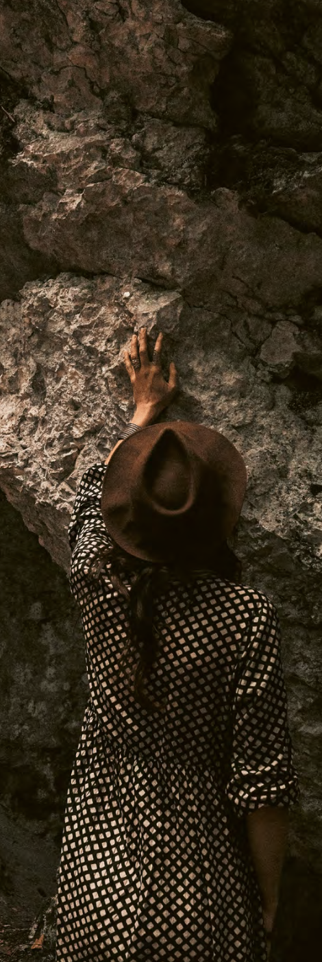 A woman wearing a brown hat and a black-and-white checkered dress reaching out to touch a rough rock wall in a cave or rocky environment.