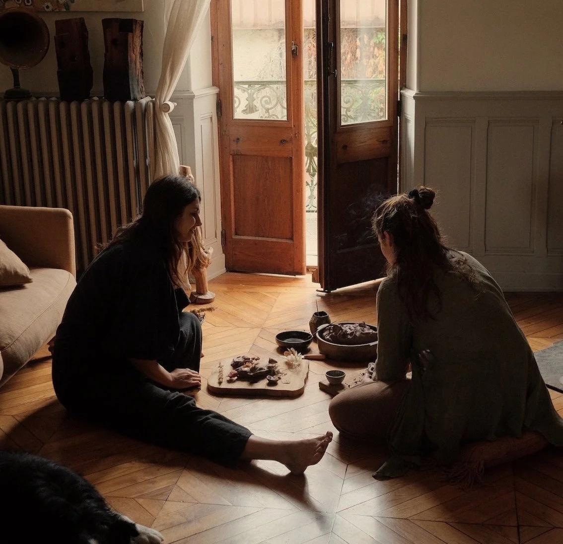 Two women sitting on the wooden floor near a door with glass panels, preparing food on a chopping board, with bowls and kitchen items around them.
