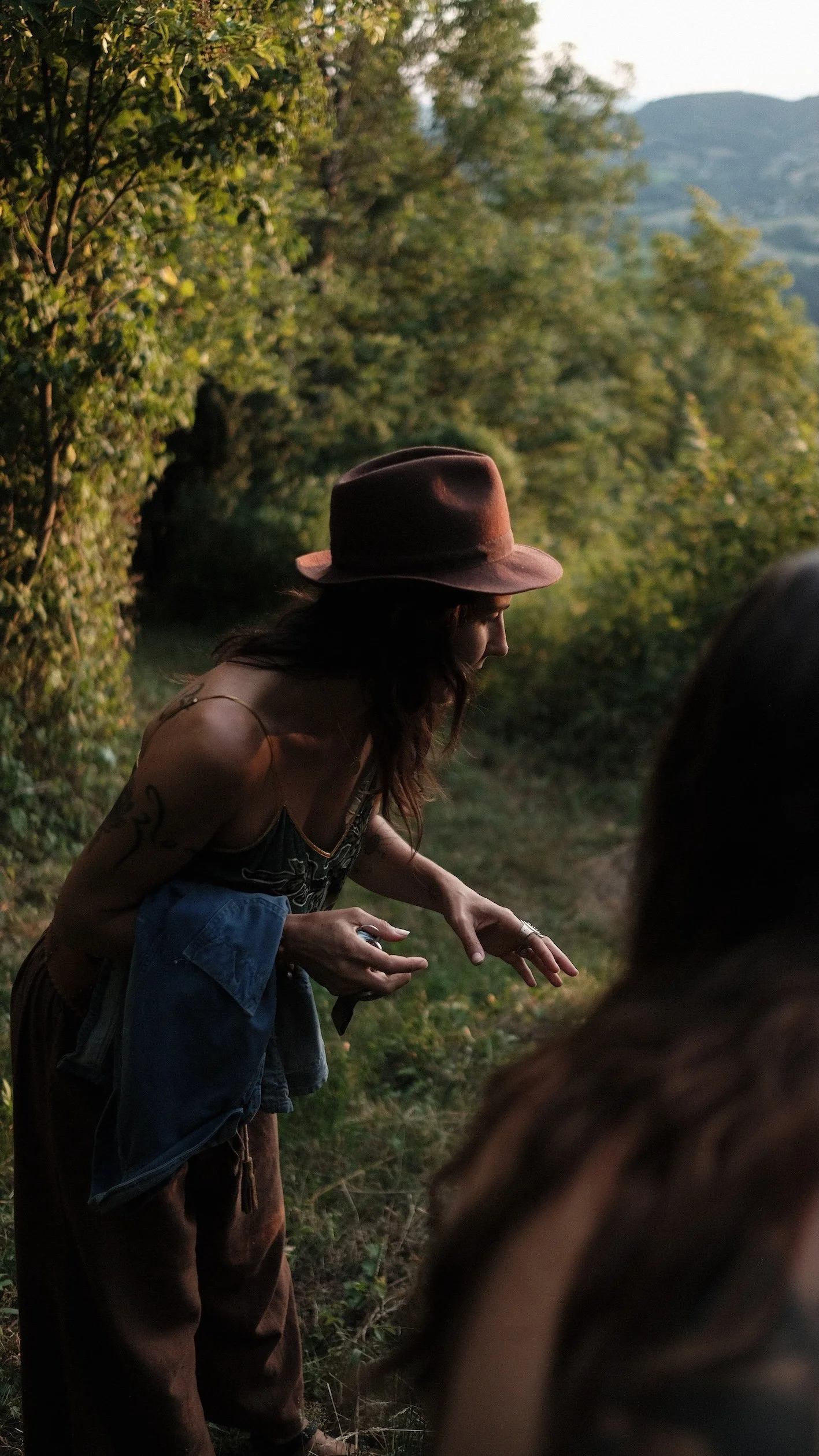 A woman wearing a brown hat, sleeveless top, and brown pants standing outdoors in a green, wooded area during sunset.