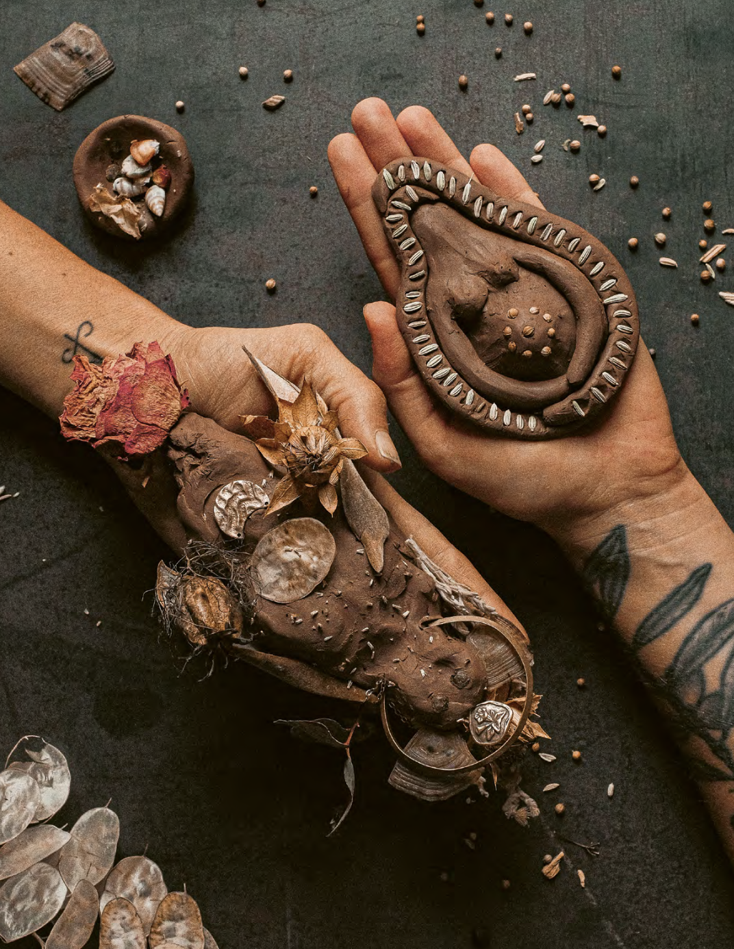 Two tattooed hands, one holding a carved wooden snake and the other a decorated wooden object, surrounded by scattered seeds and small objects on a dark surface.