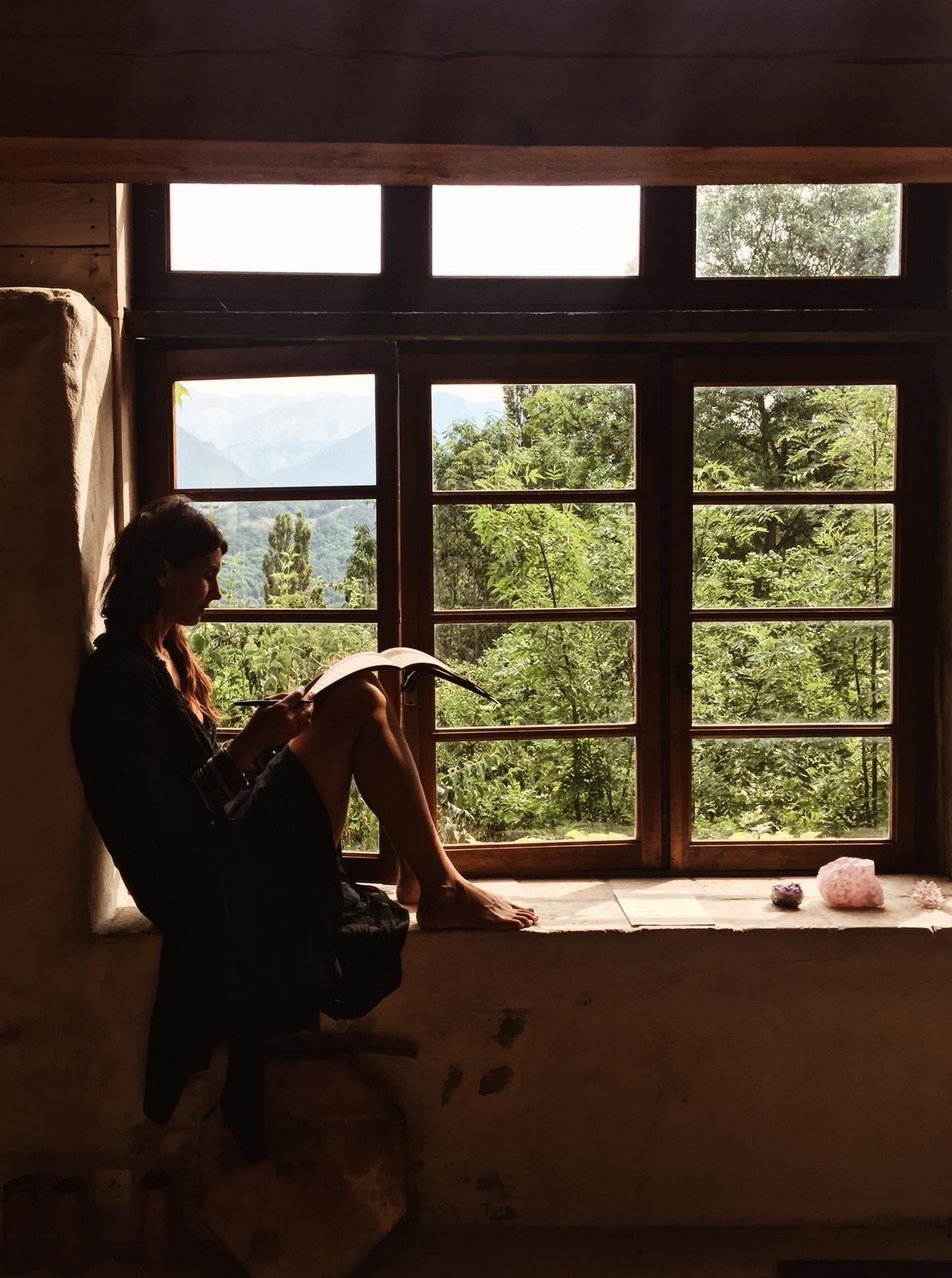 Woman sitting by a window reading a book with a view of lush green trees and mountains outside.