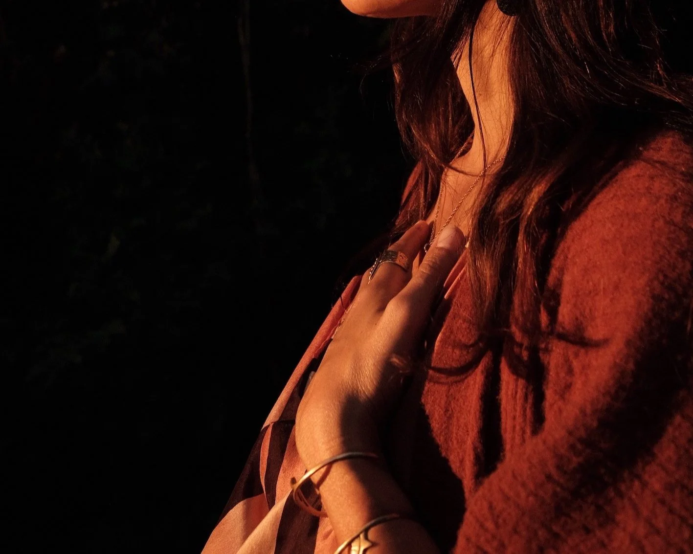 A woman with brown hair touching her chest, wearing jewelry, in a dimly lit setting with dark background.