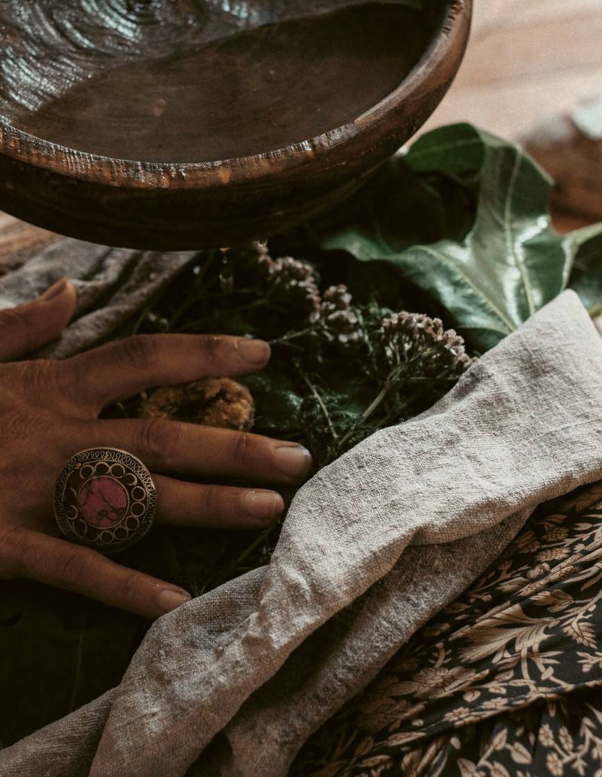 A hand with a large ornate ring on the finger is holding a dried flower stem surrounded by greenery and fabric, with a wooden bowl or dish above.