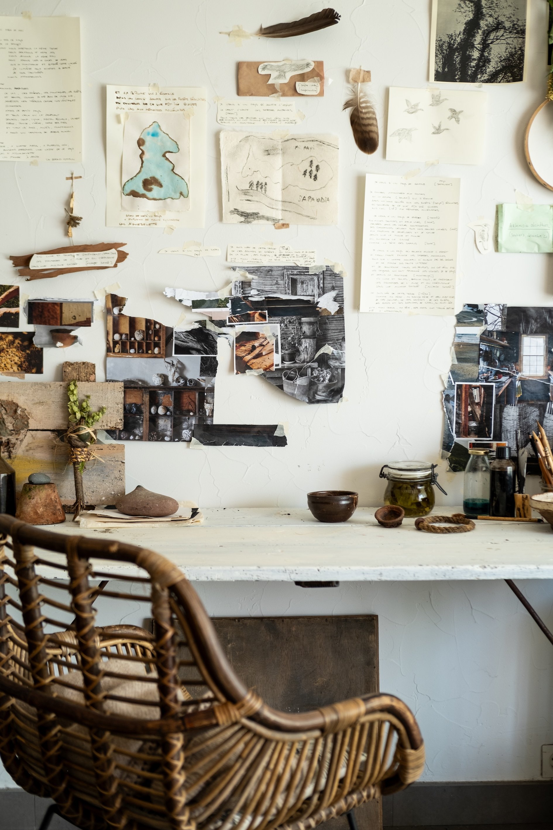 Wicker chair in front of a white wall decorated with various sketches, notes, feathers, and photographs, with a white table holding pottery and glass jars.