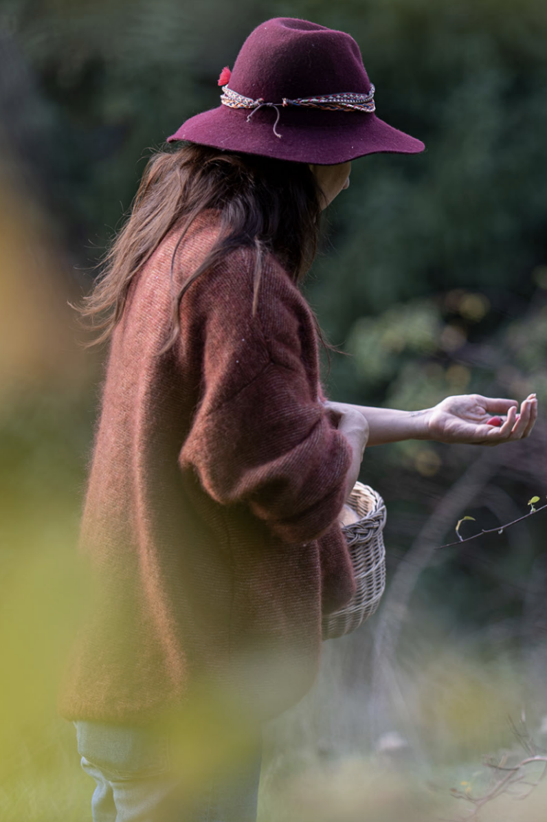 A woman wearing a burgundy hat and a rust-colored sweater standing outdoors, holding a small basket, with greenery in the background.