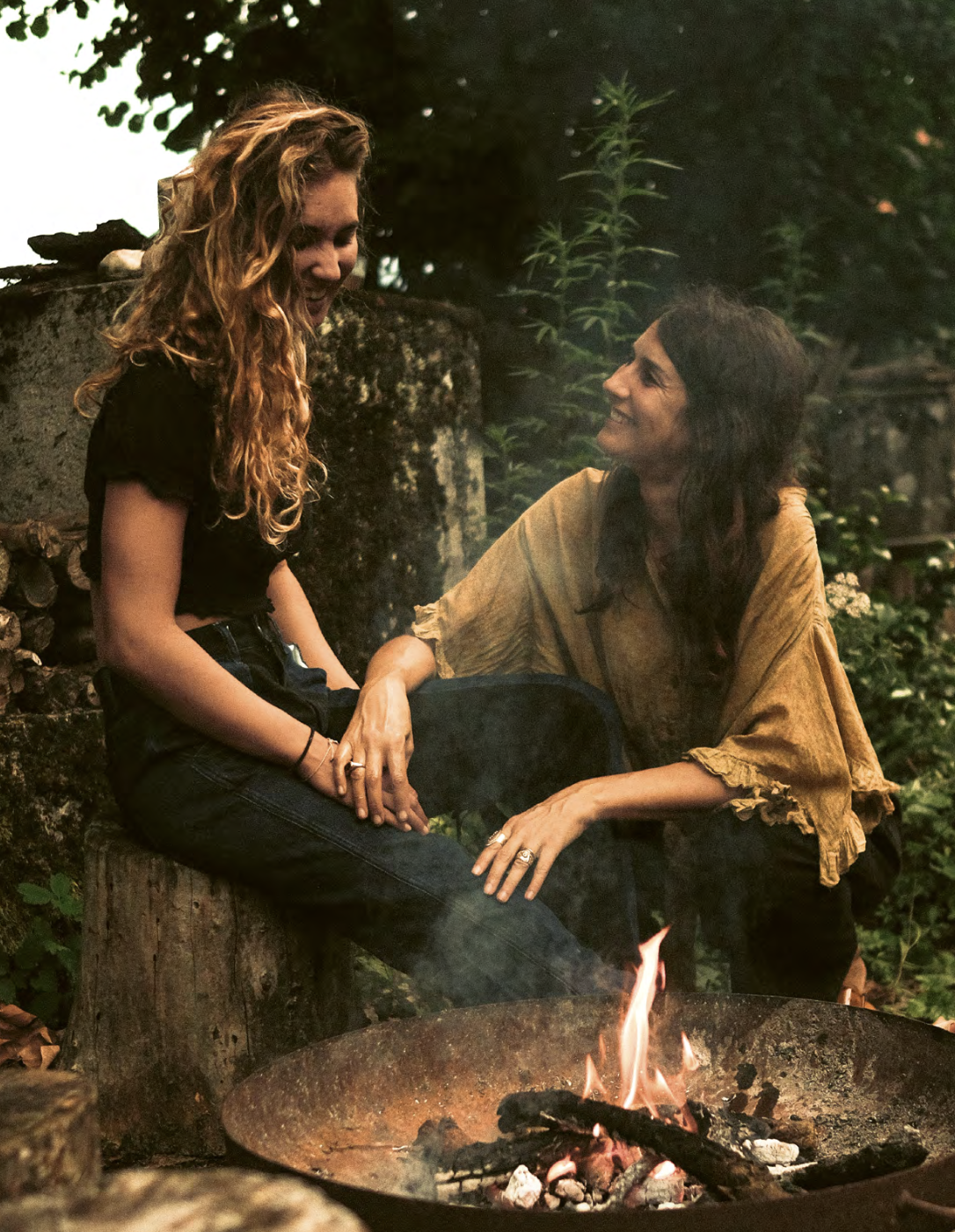 Two women sitting outdoors near a campfire, smiling and holding hands, with greenery and a stone wall in the background.