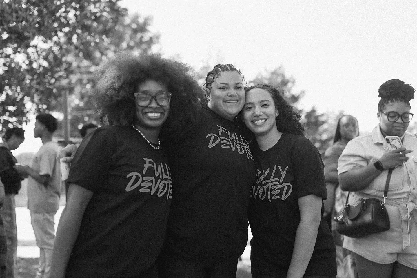 Three smiling women wearing matching black T-shirts with the words "Fully Devoted" printed on them, standing outdoors, with several people in the background.