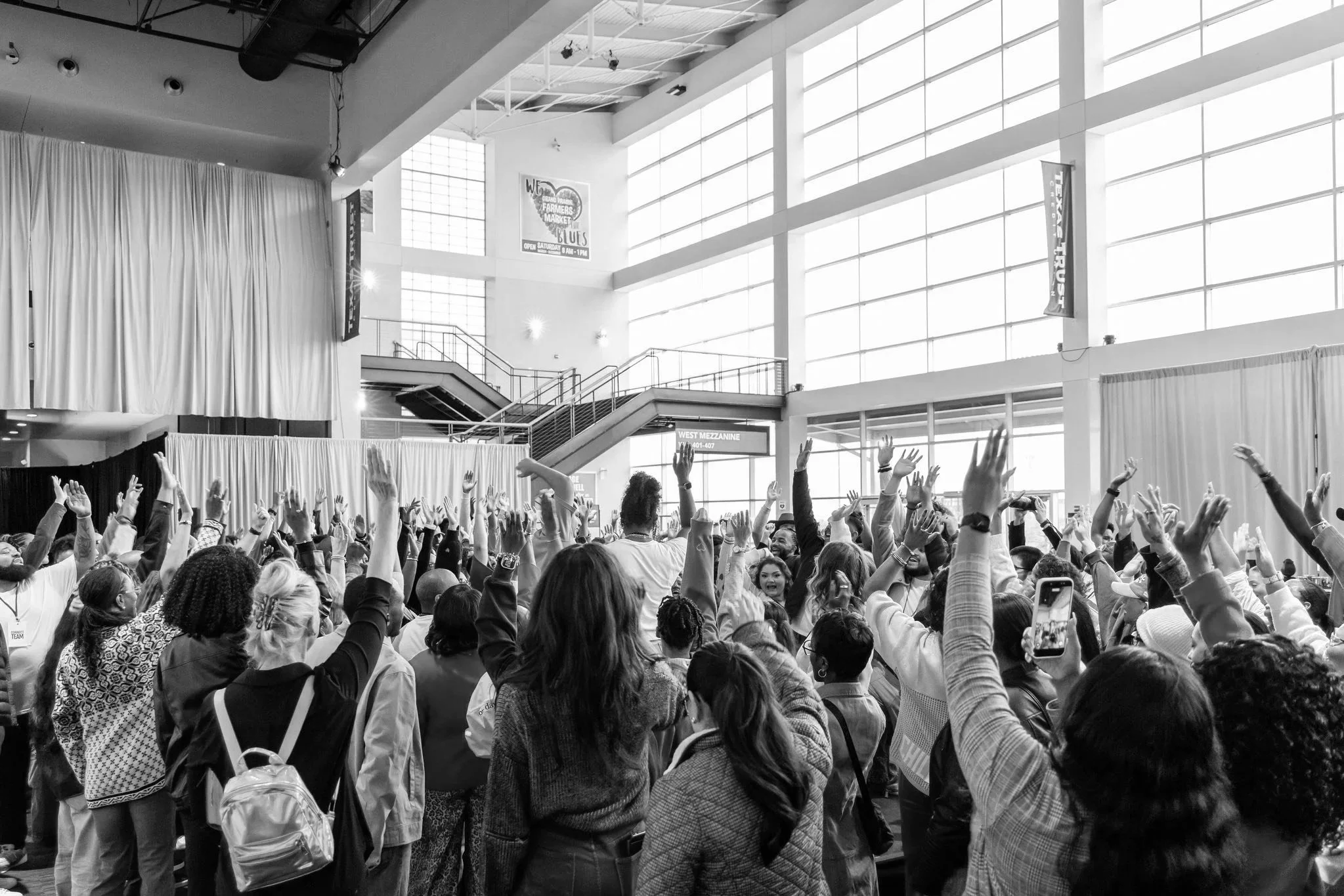 A large crowd of people at an indoor event with many raising their hands, some taking photos, in a spacious modern venue with high windows and stage curtain.
