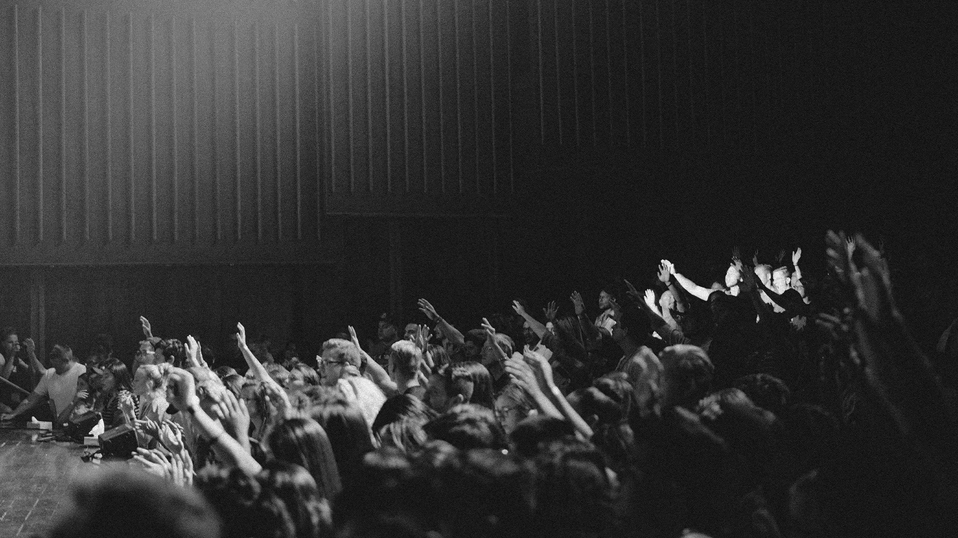 Black and white photo of a large audience in a concert hall, with many people raising their hands.