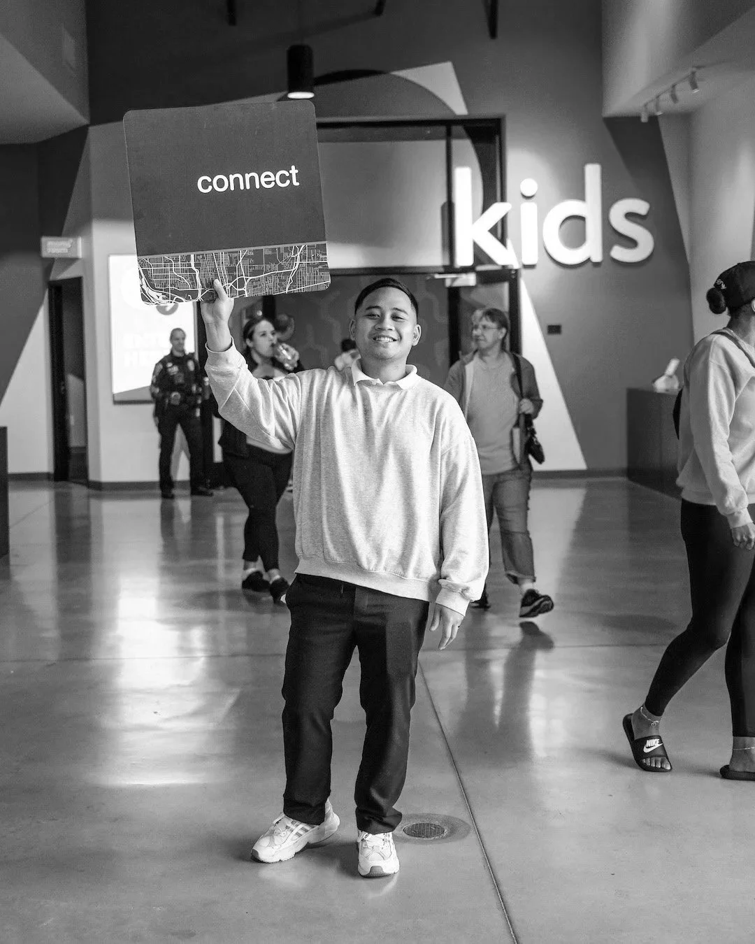 A smiling boy in a light hoodie and dark pants holding a sign that says "connect" inside a shopping mall or large indoor space with other people walking by.