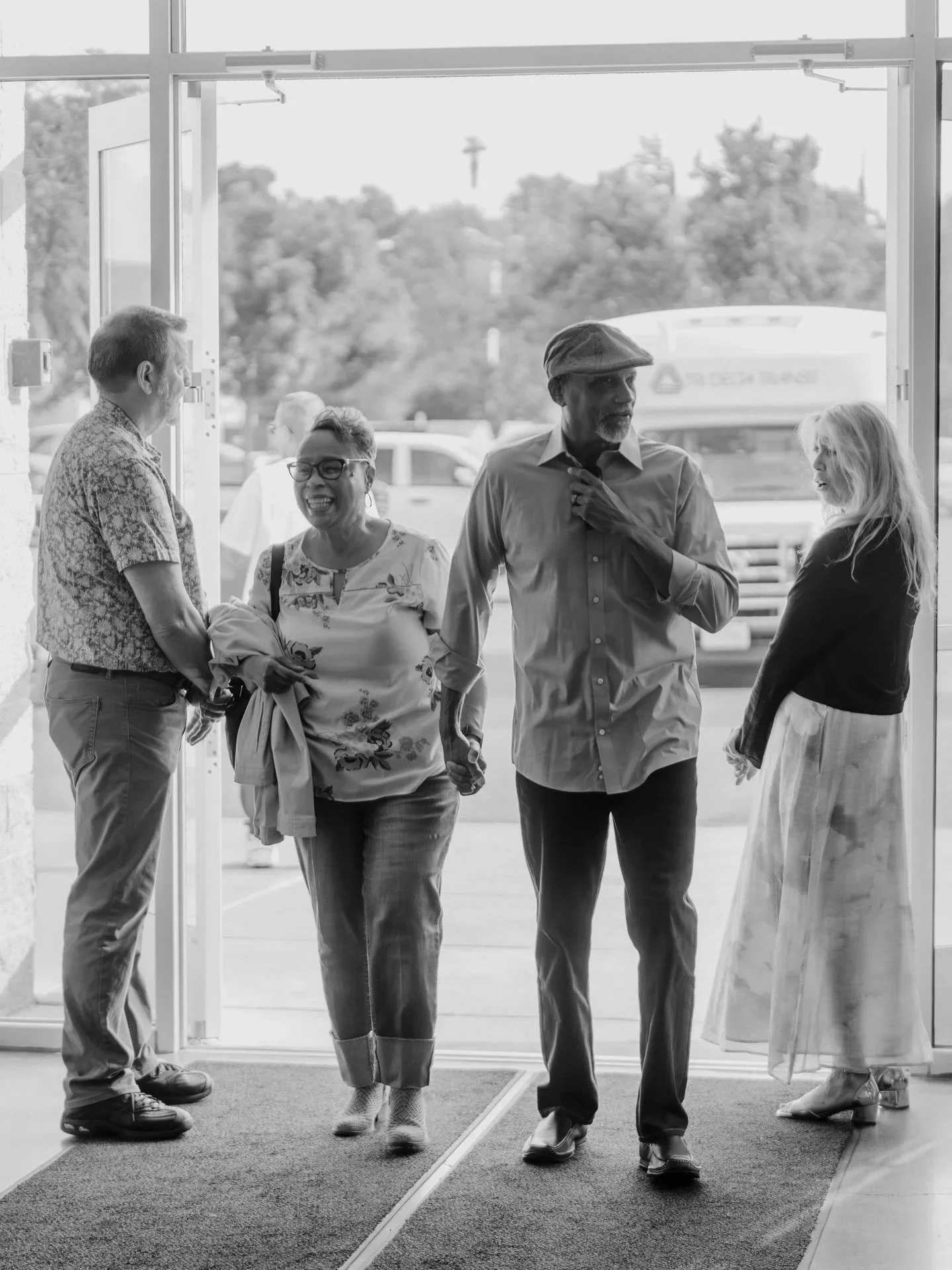 Four people standing at the entrance of a building, smiling and chatting. The door is open, revealing a parking lot outside. The individuals include two men and two women, dressed casually, appearing to enjoy a friendly conversation.