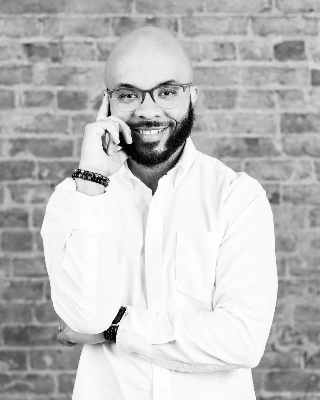 Portrait of a smiling man with glasses, a beard, and a bald head, wearing a white shirt, standing in front of a brick wall.