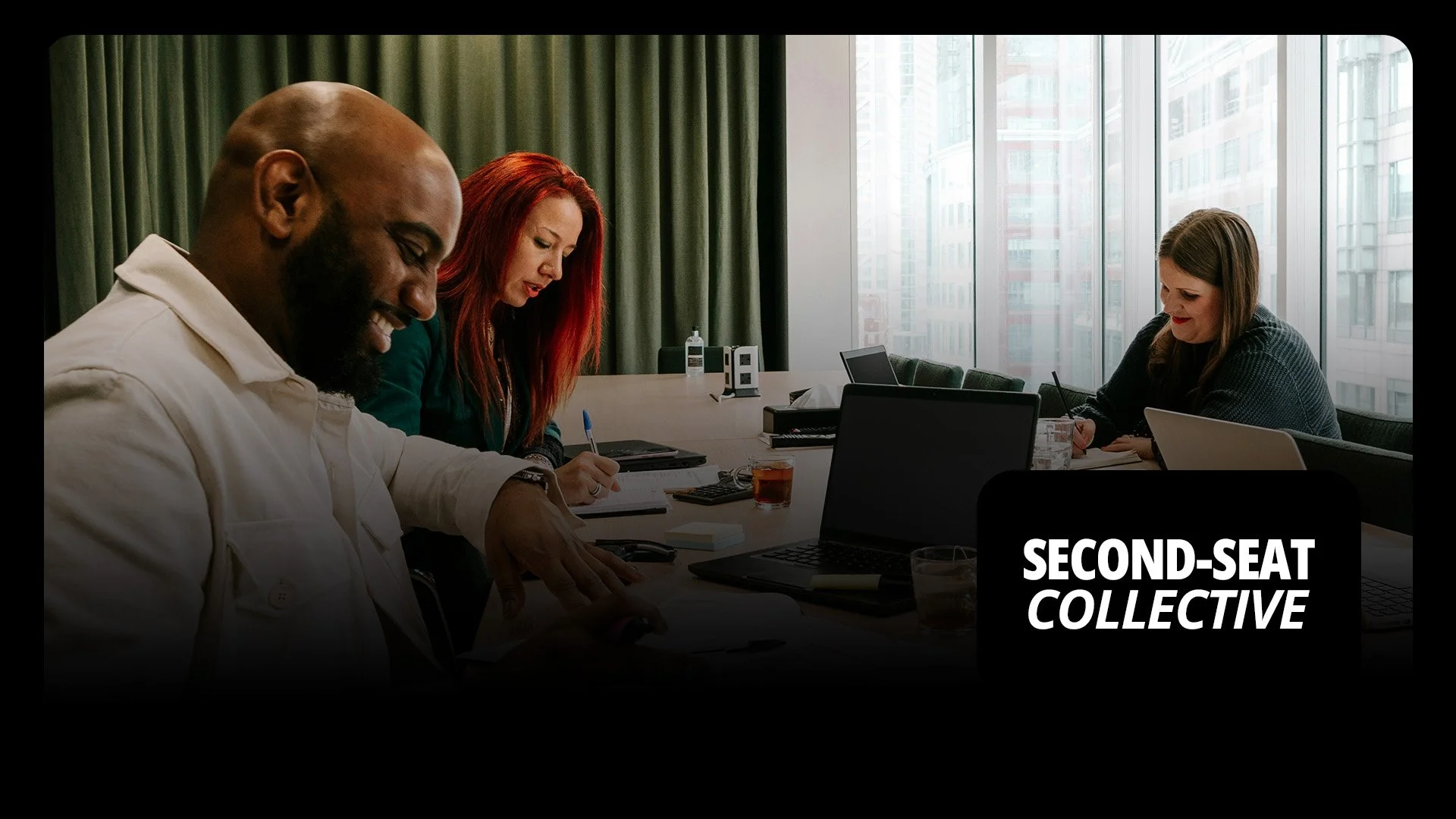 Three people sitting at a table in a conference room with large windows, working on laptops and taking notes. The scene suggests a professional meeting or collaboration.