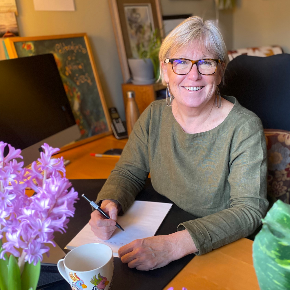 Helen smiling, sitting at a desk, writing on a notepad with a pen. There are purple flowers in a vase, a coffee mug, and a computer on the desk. In the background, framed artwork and a plant are visible.