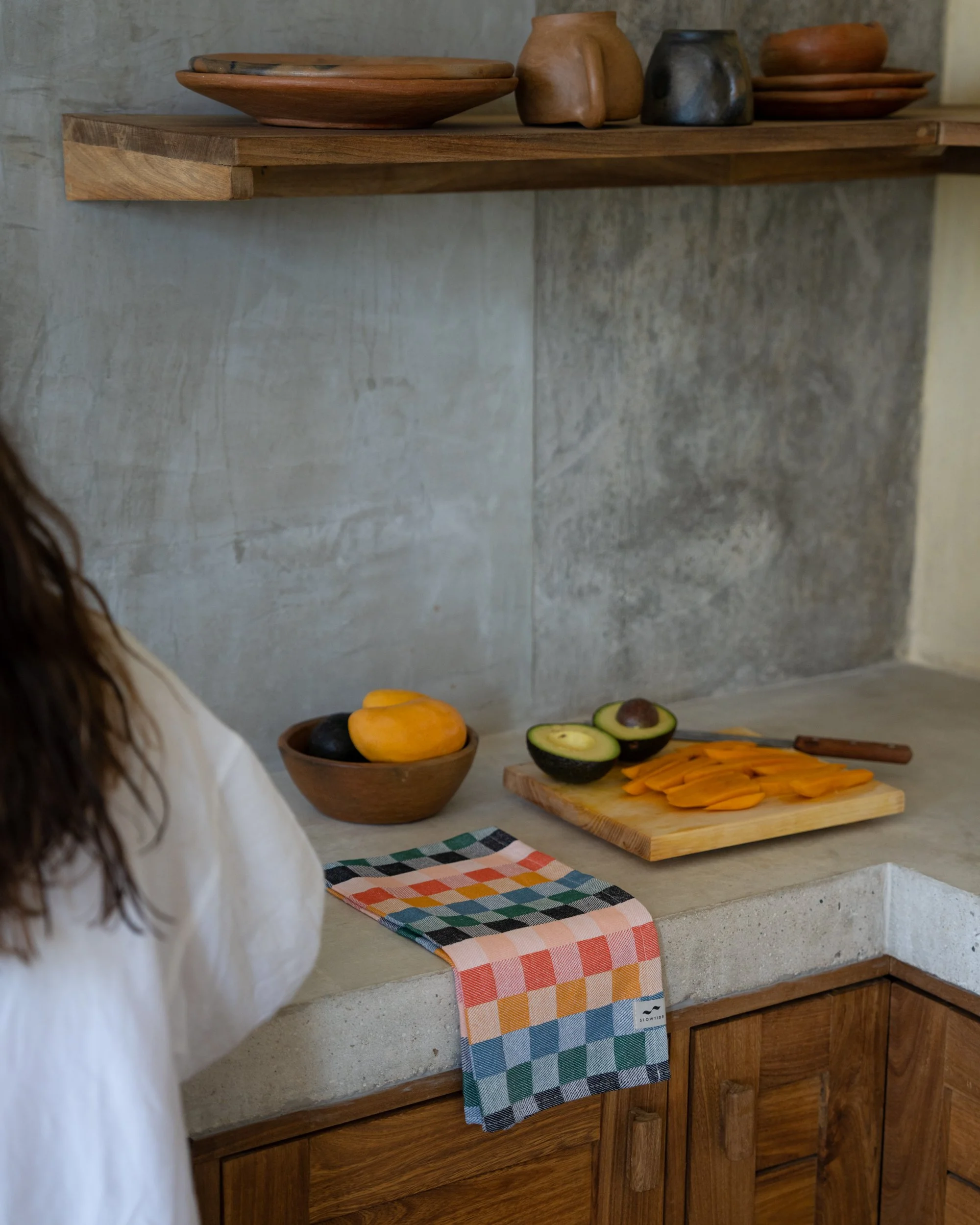Kitchen countertop with sliced mango, avocado halves, and a bowl of whole avocados, with a colorful checkered cloth and wooden cabinets.