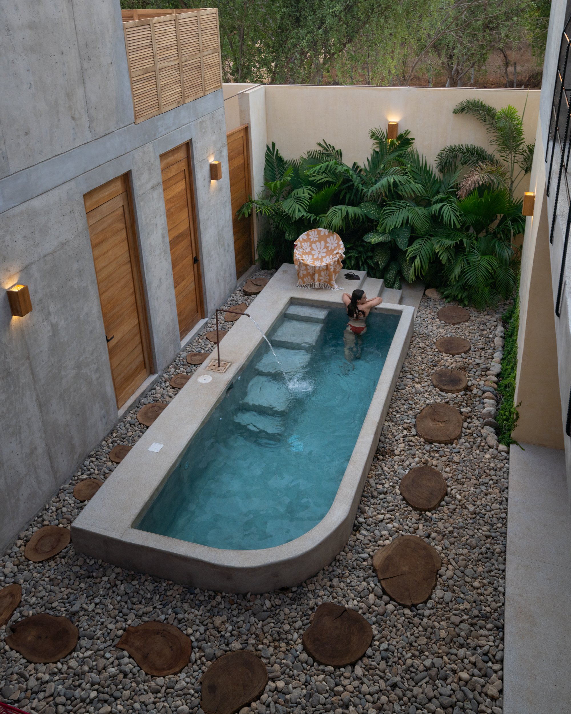 A small rectangular outdoor pool with a woman swimming and relaxing, surrounded by tropical plants, rock details, and modern wall-mounted lights.