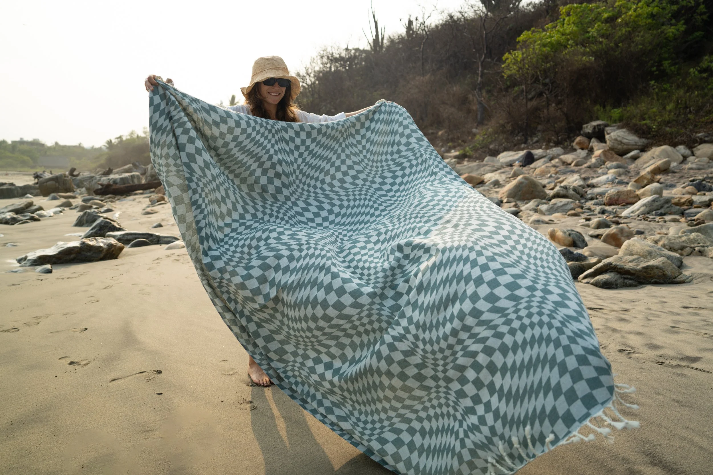Woman on a sandy beach holding a large checkered towel, wearing a sun hat and sunglasses, with rocks and trees in the background.