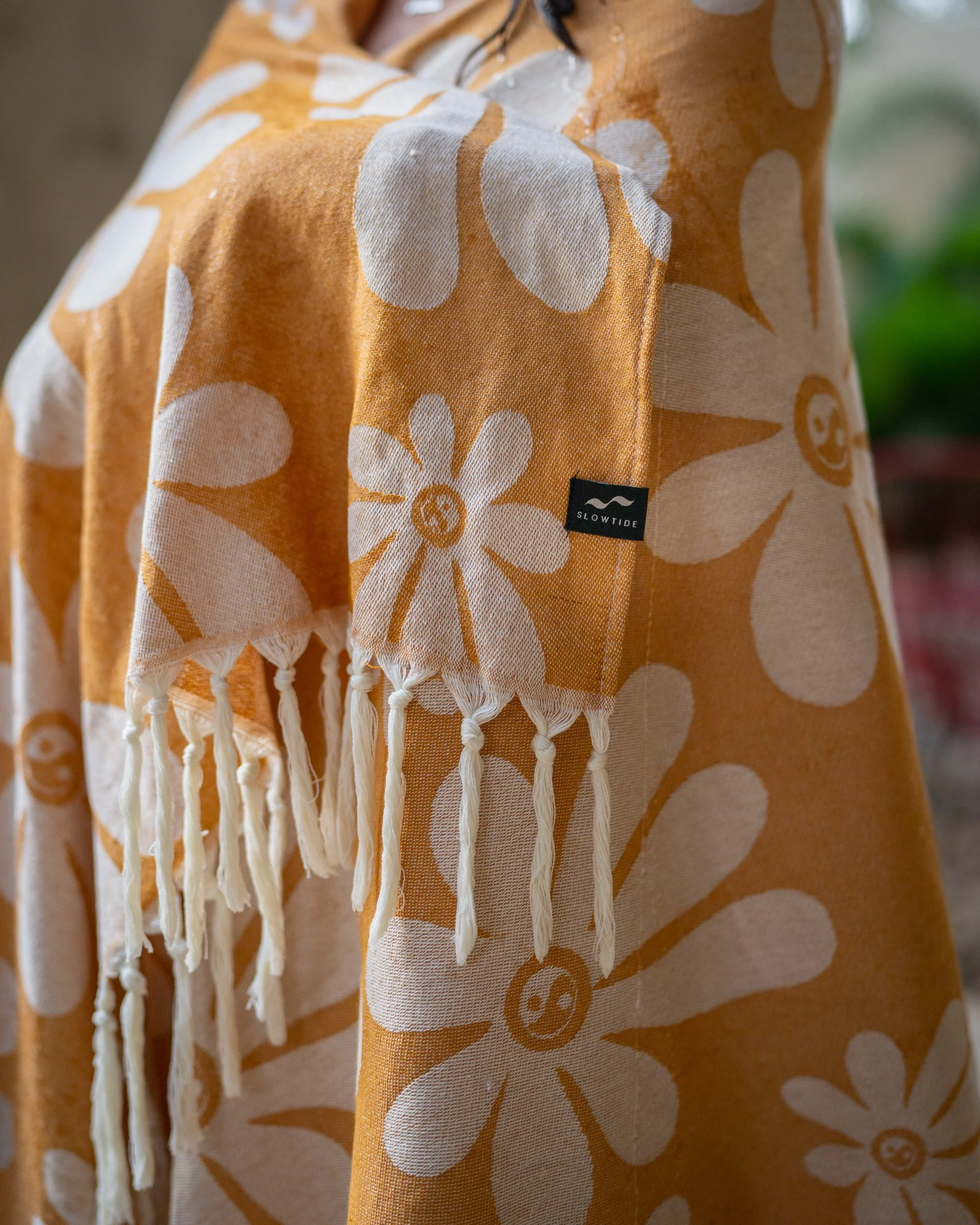 A close-up of an orange floral patterned fabric with fringes, featuring a black label that reads 'SLOWTIDE'.