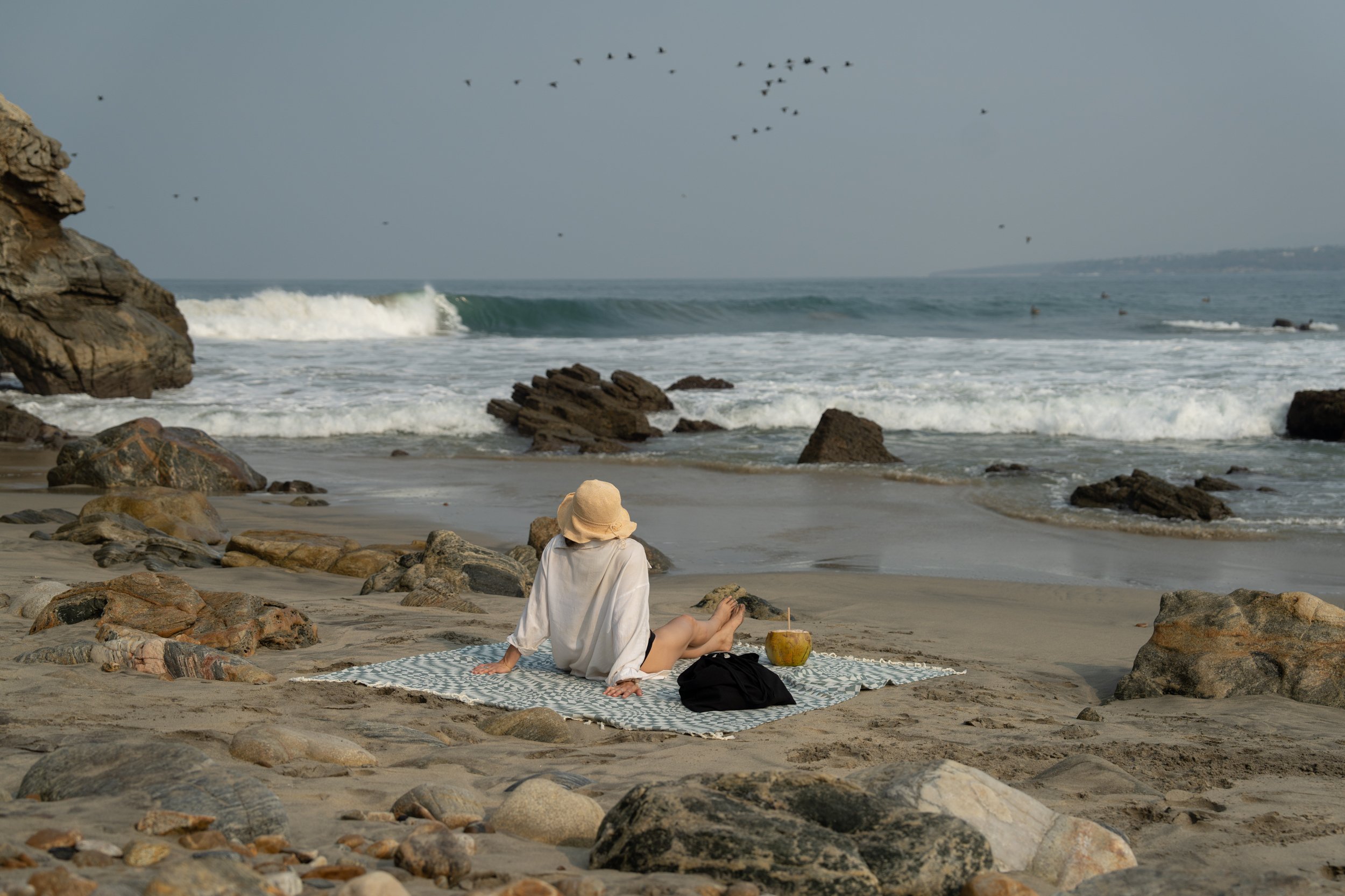 A person sitting on a beach blanket facing the ocean, wearing a hat and relaxing as waves crash onto rocks and shore in the background.