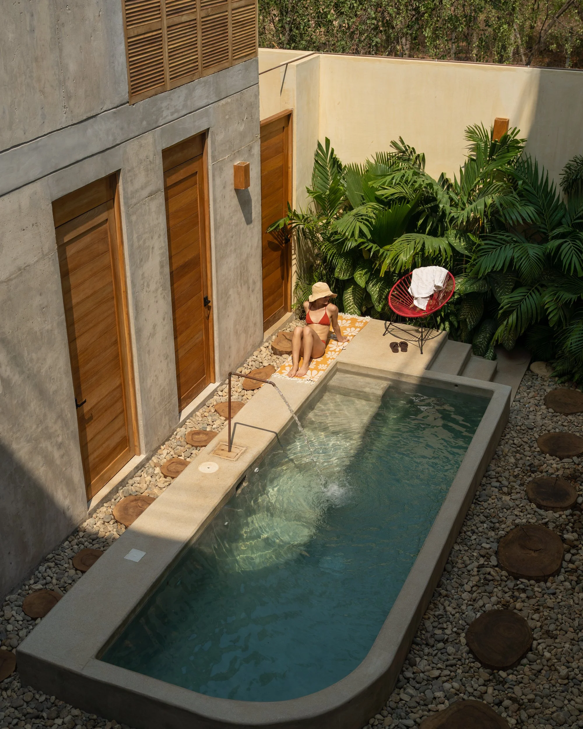 A woman relaxing on a towel next to a small rectangular swimming pool, surrounded by rocks and lush green plants, with a modern house wall in the background.