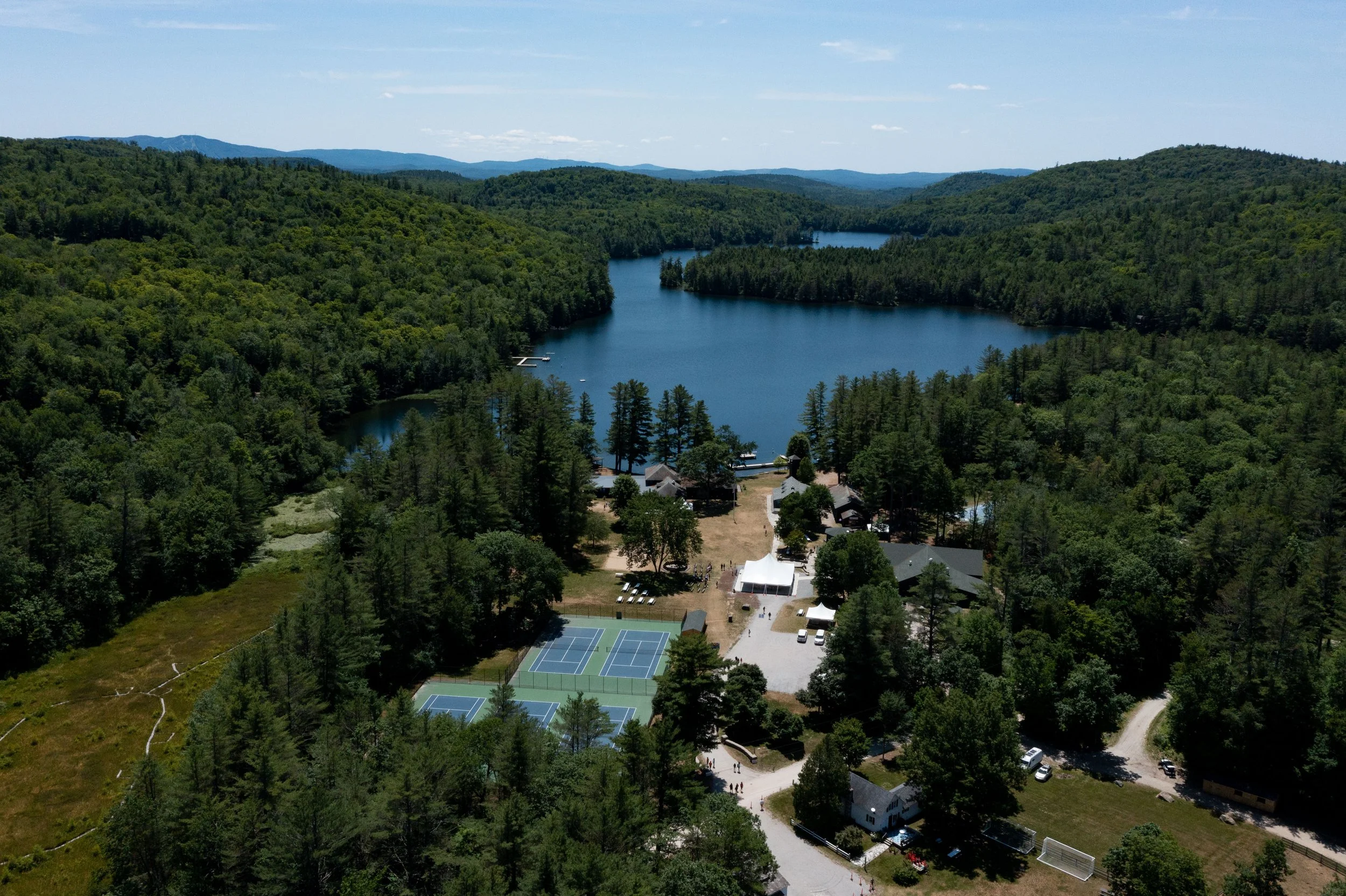 Aerial view of a lakeside area surrounded by dense green trees with tennis courts, buildings, and a large body of water in the background.
