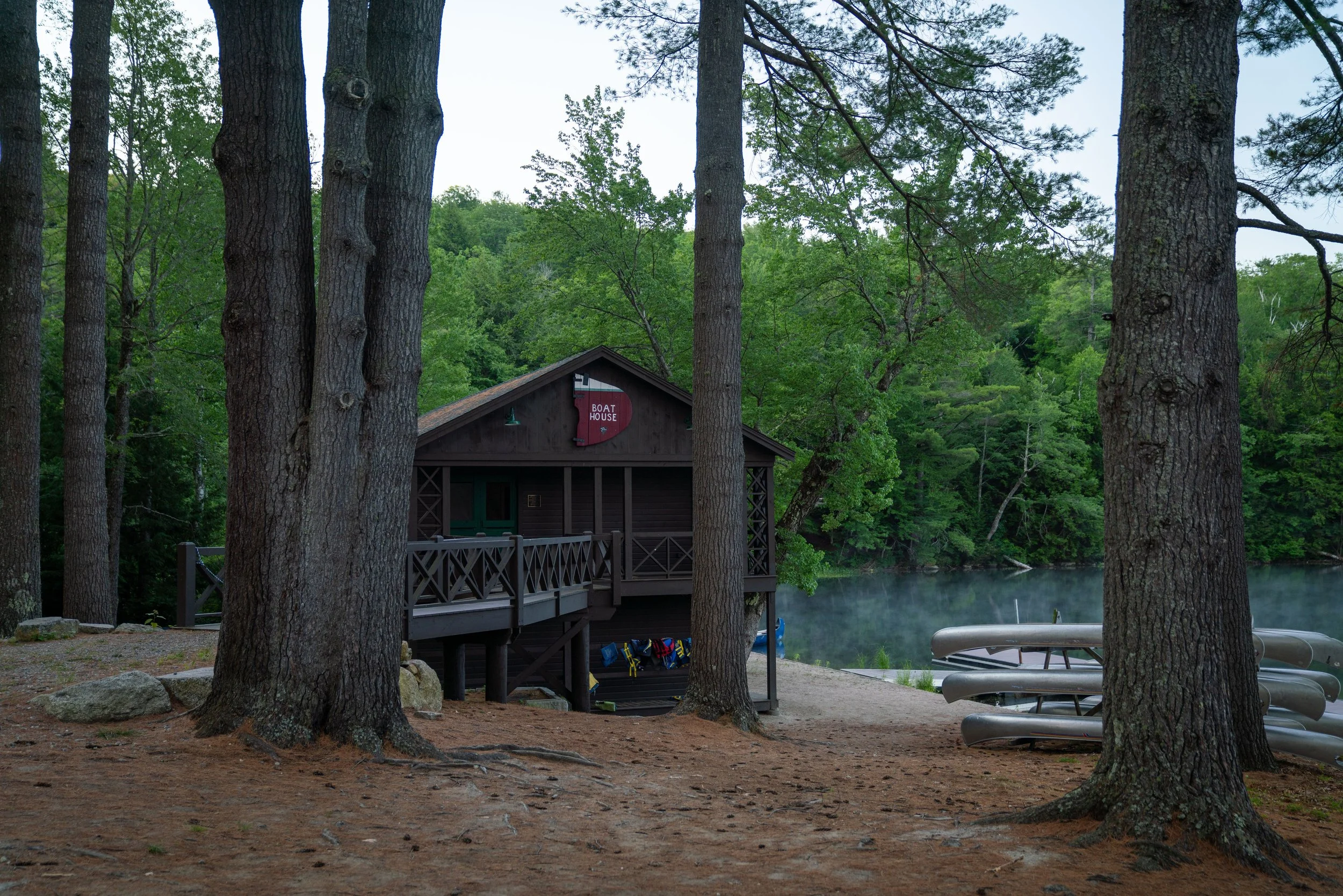 A wooden boathouse labeled 'Boat House' located by a lake surrounded by green trees, with multiple canoes stacked nearby and a dock extending over the water.