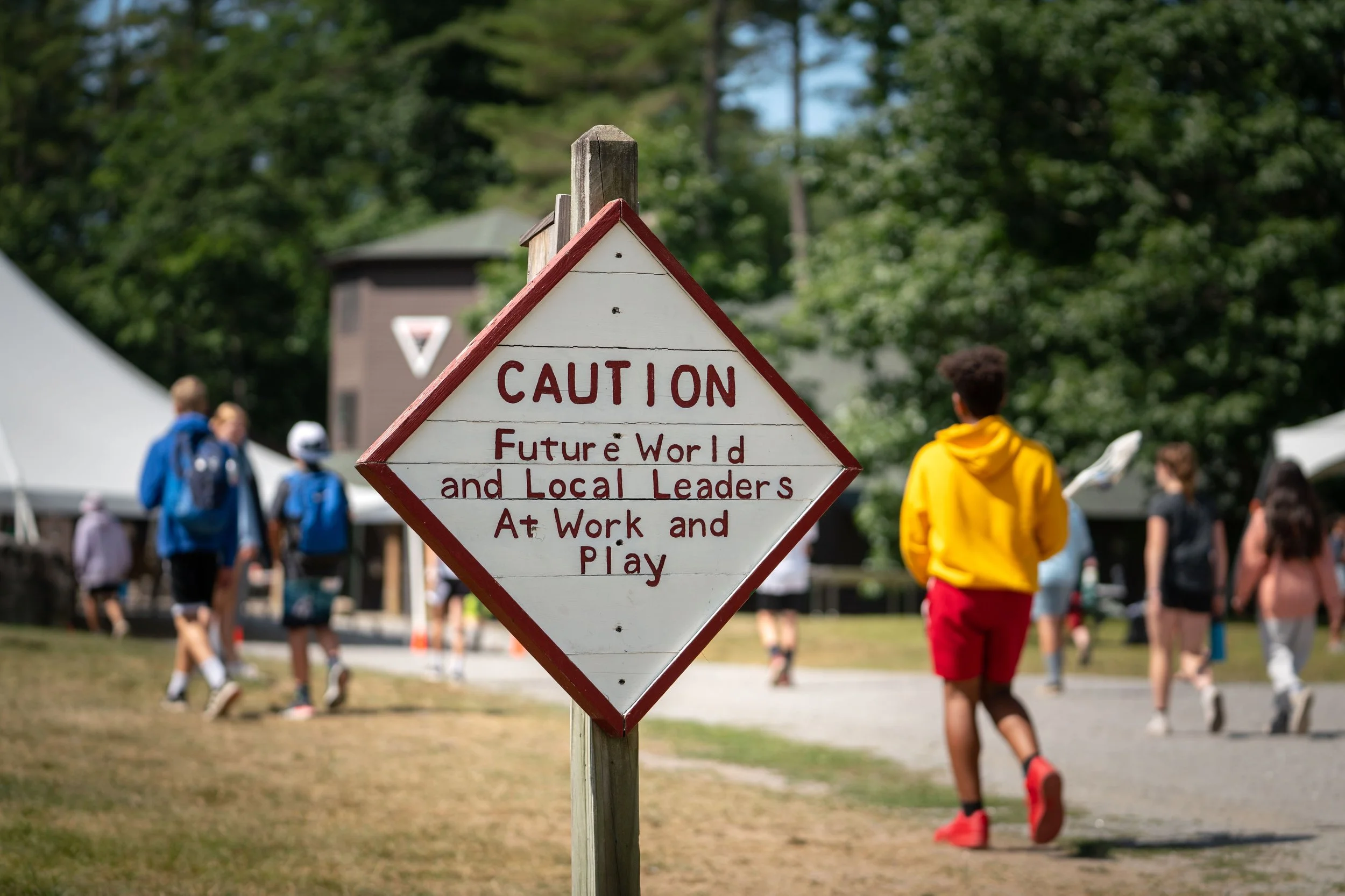 A sign that reads 'CAUTION Future World and Local Leaders At Work and Play' at an outdoor event with children walking and playing in the background.
