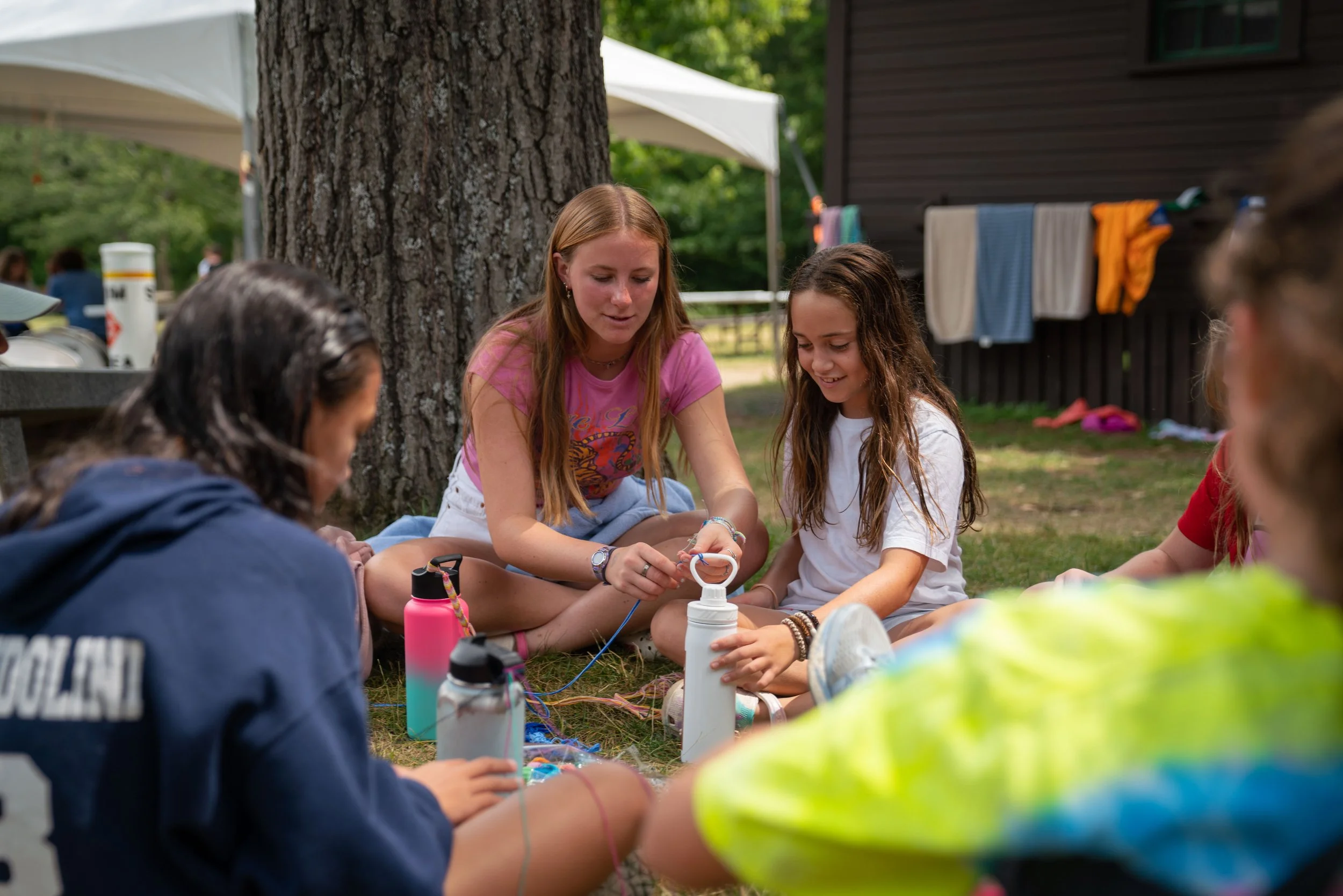 Group of girls sitting on grass outdoors, gathered around a white water bottle, engaging in a craft activity, with trees and a cabin in the background.