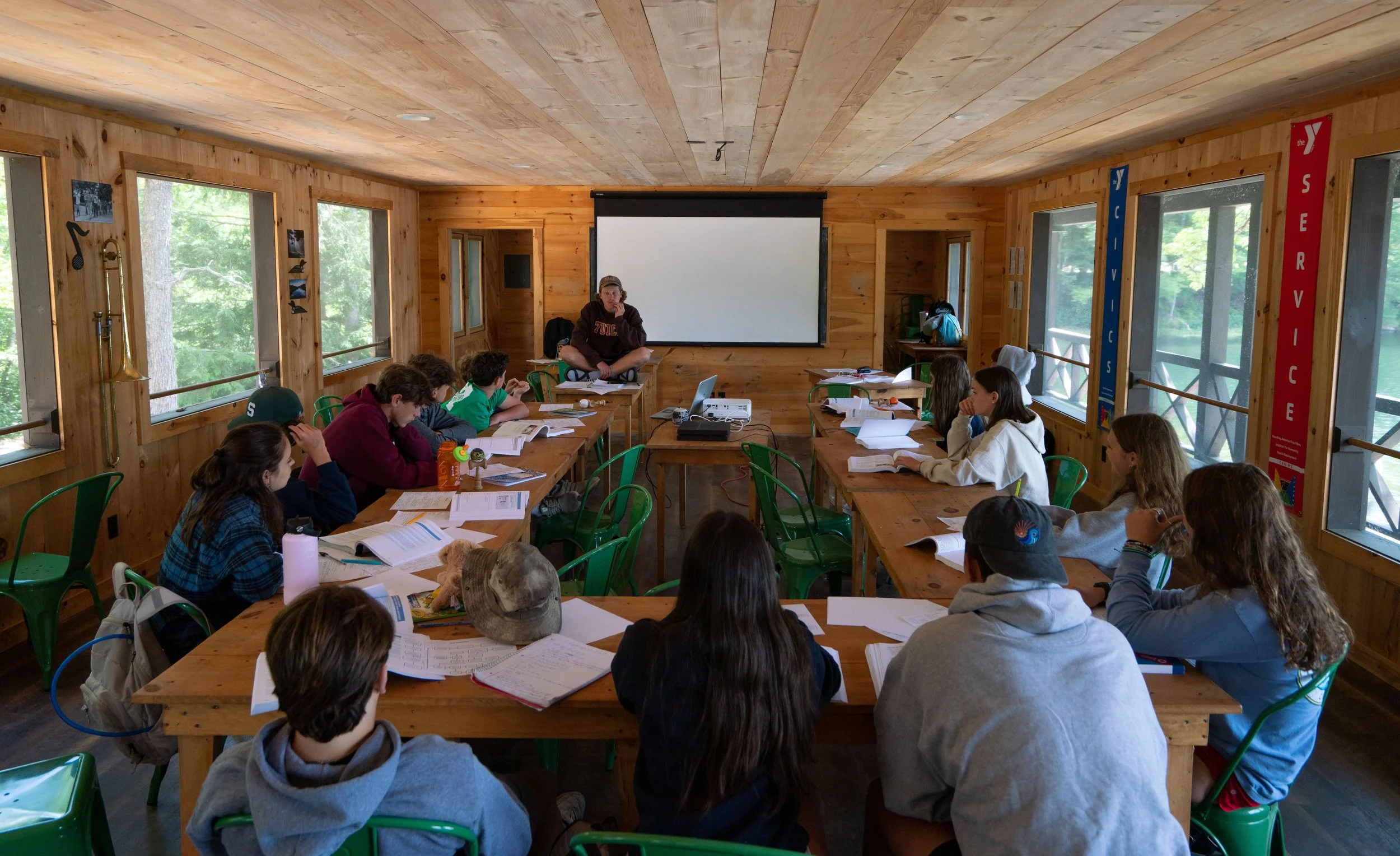 A classroom with students sitting around wooden tables, facing a teacher at the front. The room has wood-paneled walls and large windows with views of trees outside. There is a white projection screen behind the teacher, and the students have open bo