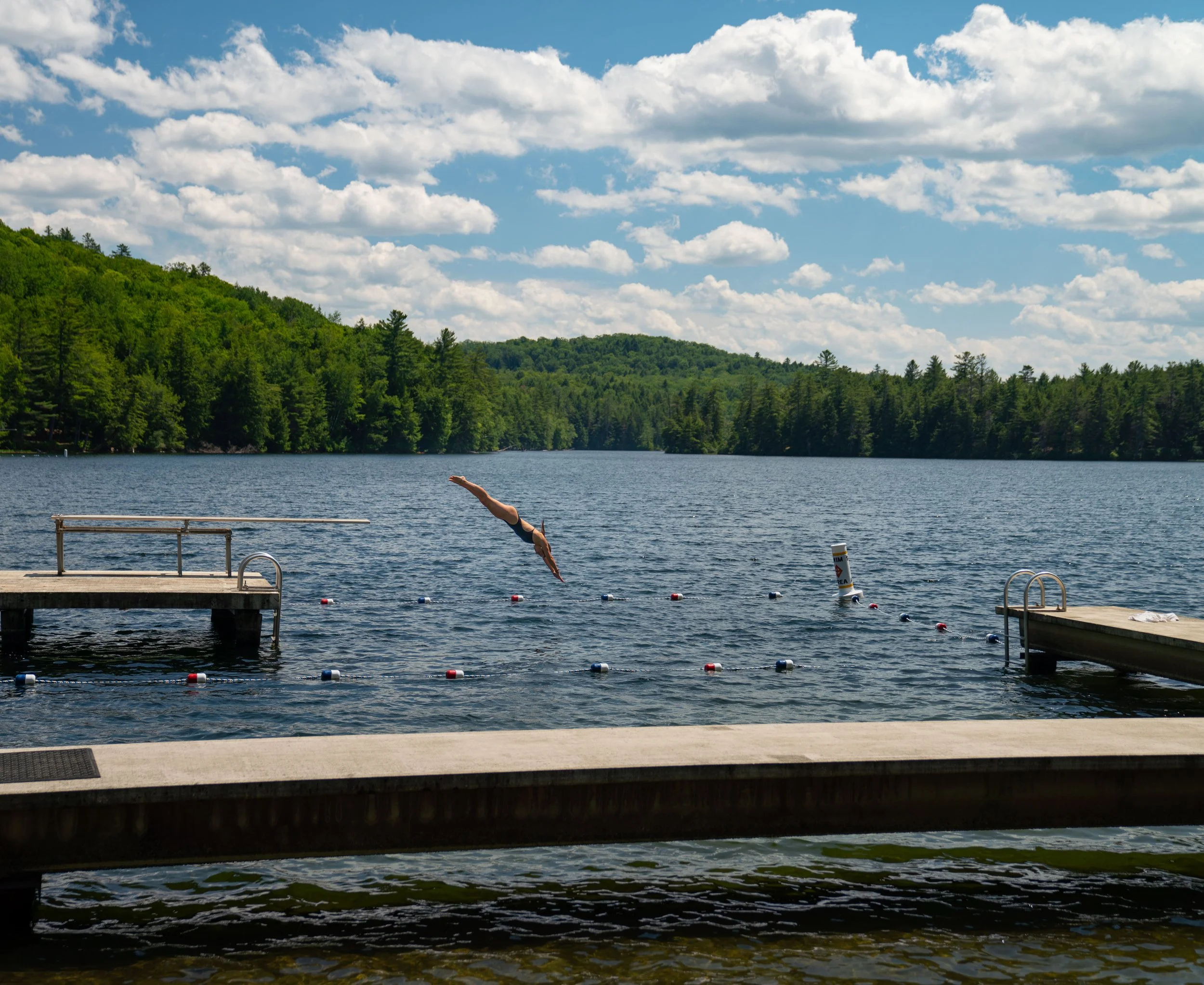 A person diving into a lake from a dock on a sunny day with a forested shoreline and partly cloudy sky.