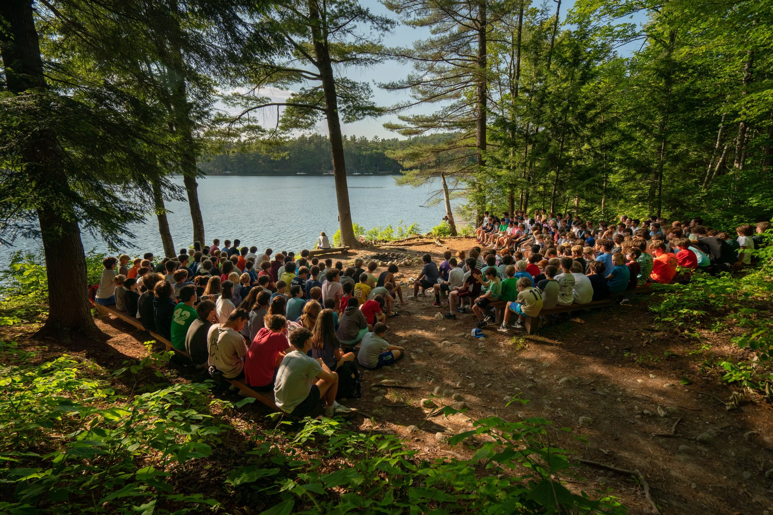 Large group of children and teenagers sitting on benches and on ground in a forested area by a lake, attending an outdoor event or camp activity on a sunny day.