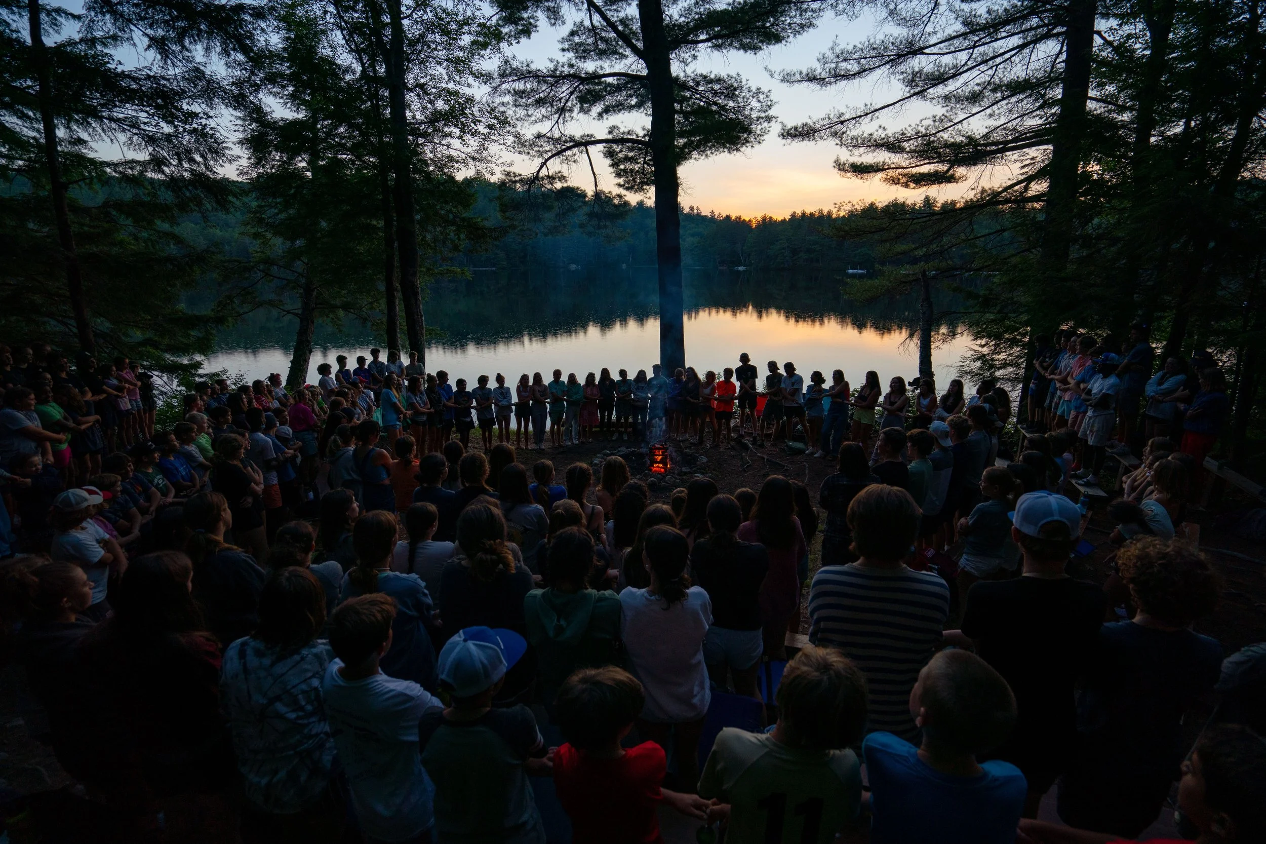 A large group of children and adults gathered around a campfire at sunset near a lake, surrounded by trees.