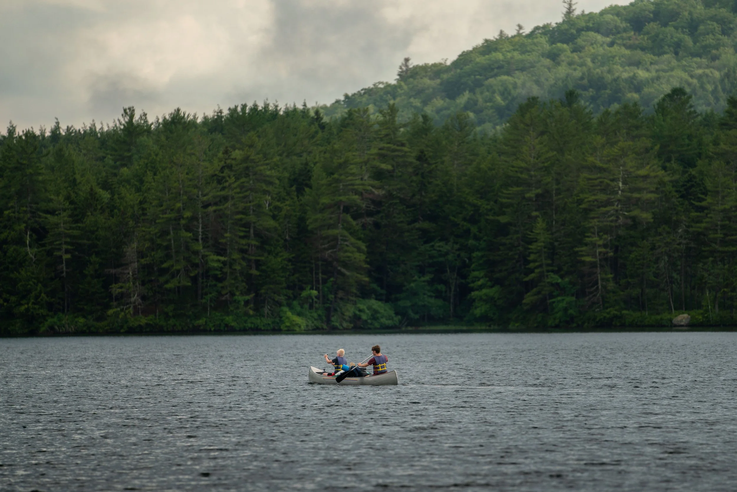 Two people in a canoe on a lake, surrounded by green trees under a cloudy sky.