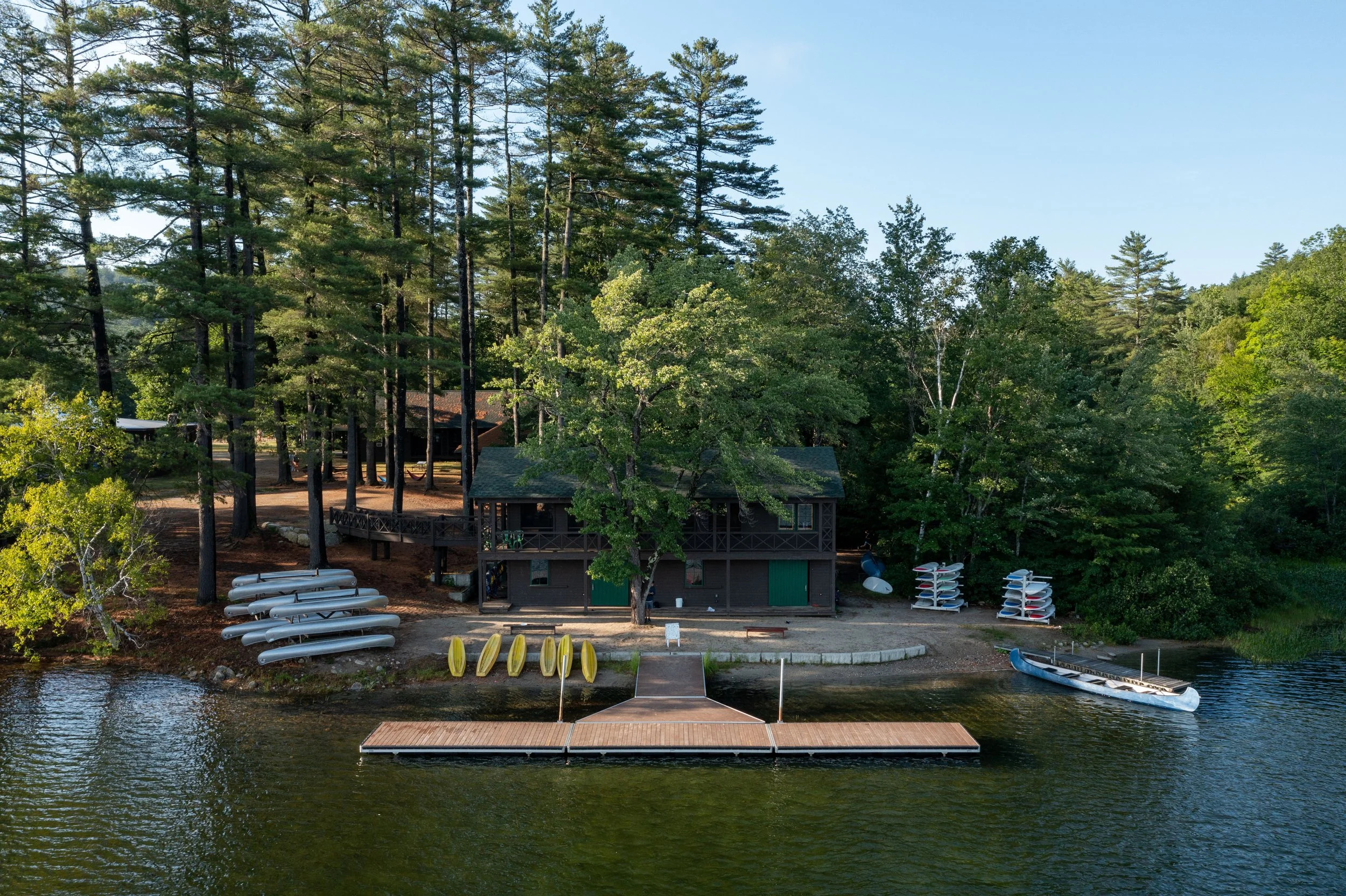 A lakeside cabin surrounded by tall pine trees, with canoes, kayaks, and paddleboards stored outside, a dock extending into the water, and a boat nearby, in a peaceful, wooded setting.