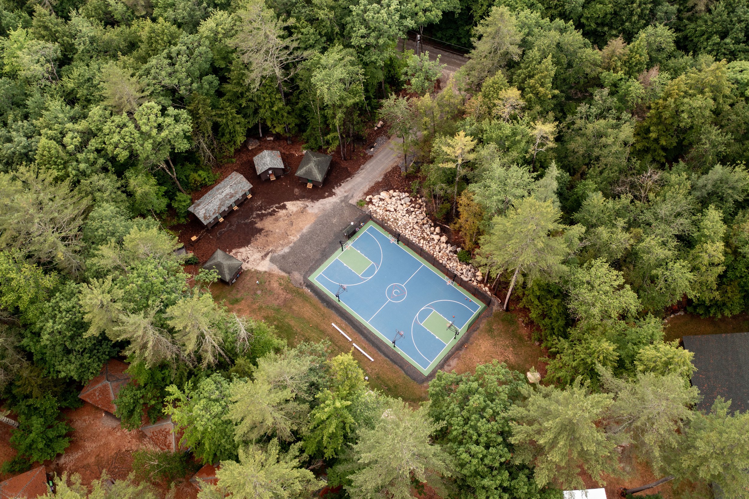 An aerial view of a blue basketball court surrounded by green forest, with four small wooden cabins nearby and a dirt pathway leading into the woods.