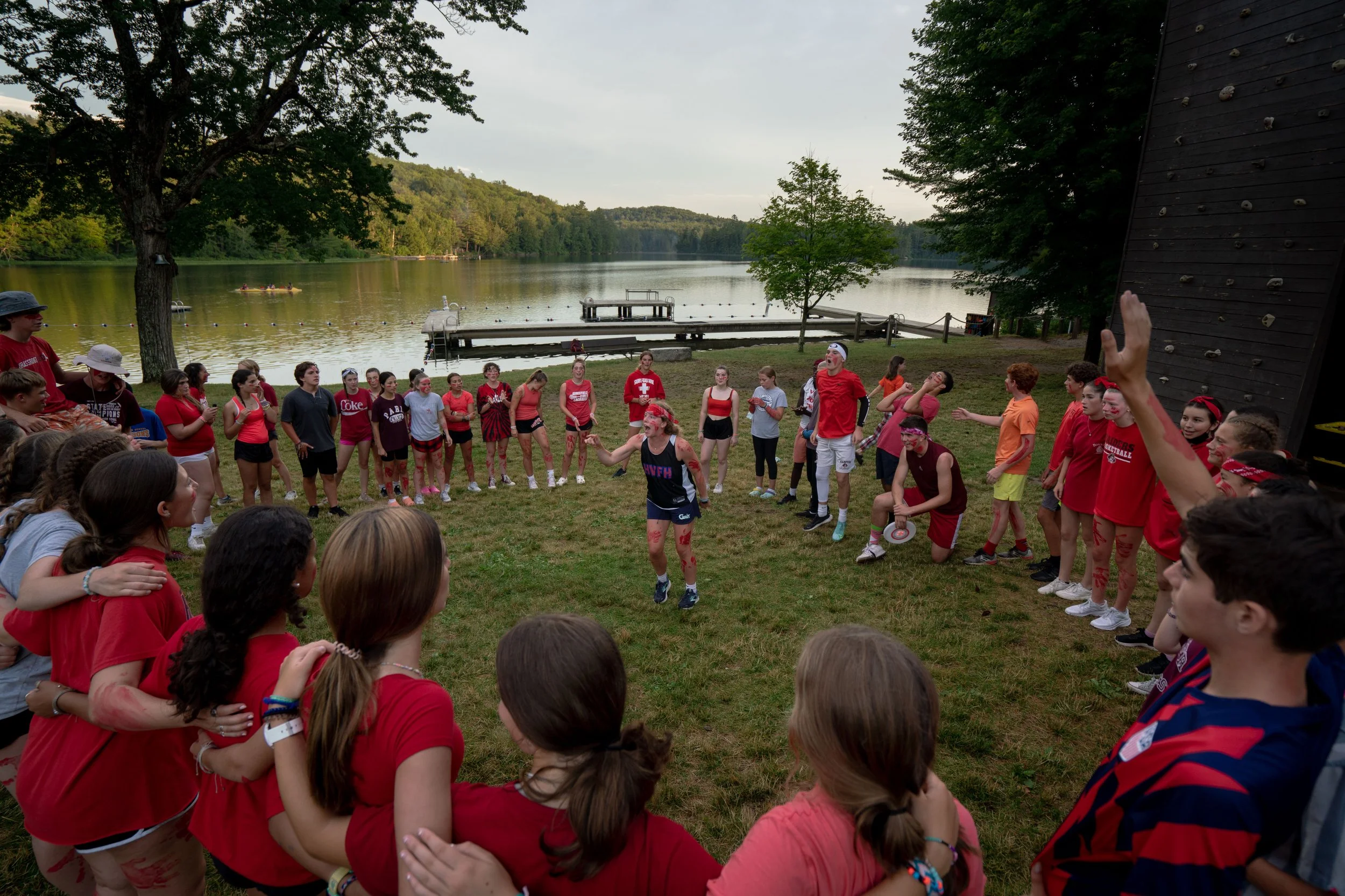 Group of people gathered outside near a lake, participating in a team-building activity or game, with trees and water in the background.