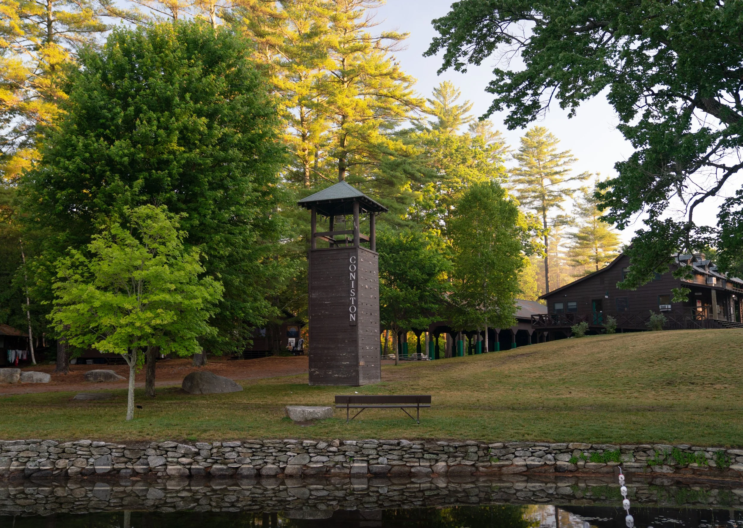 A park scene with a wooden bell tower labeled 'Coniston', green trees, grassy terrain, a stone-lined waterway in the foreground, and a brown building in the background, illuminated by soft sunlight.