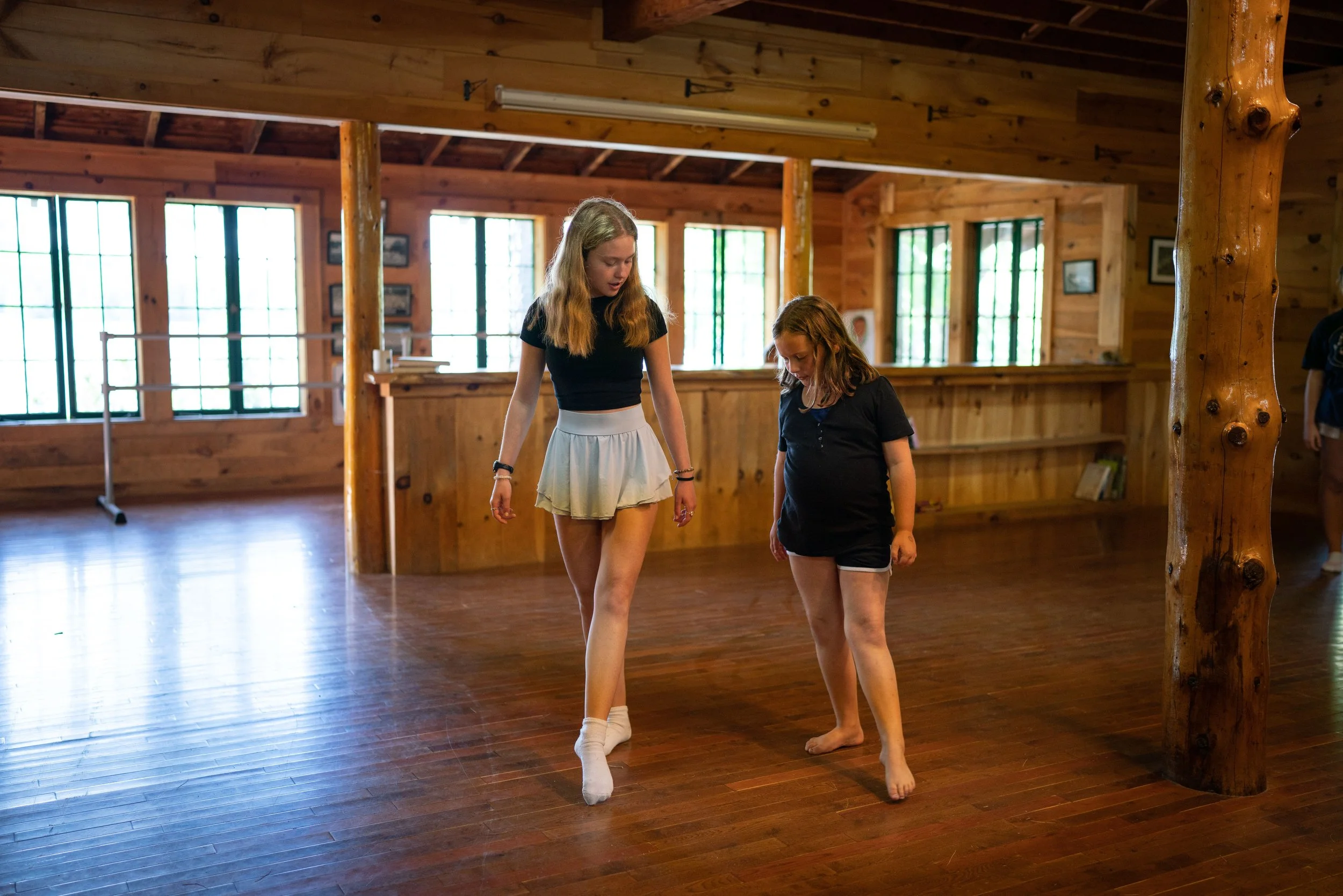 Two girls in a wooden dance studio, one with long blonde hair wearing a black shirt and white skirt, standing on her toes, and the other with red hair wearing a black shirt and shorts, looking down at her feet.