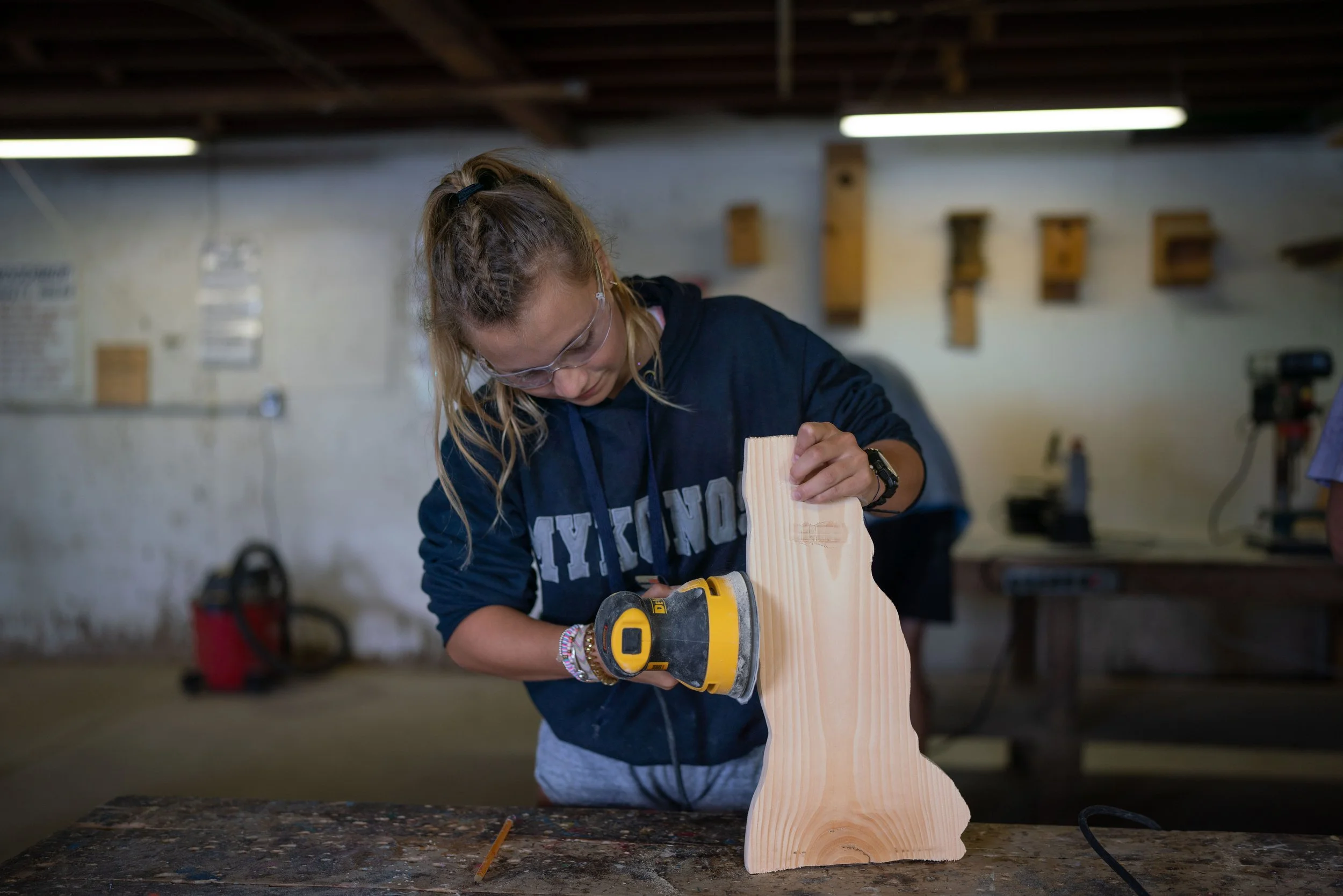 A girl wearing safety goggles using a yellow power sander on a piece of wood in a woodworking workshop.