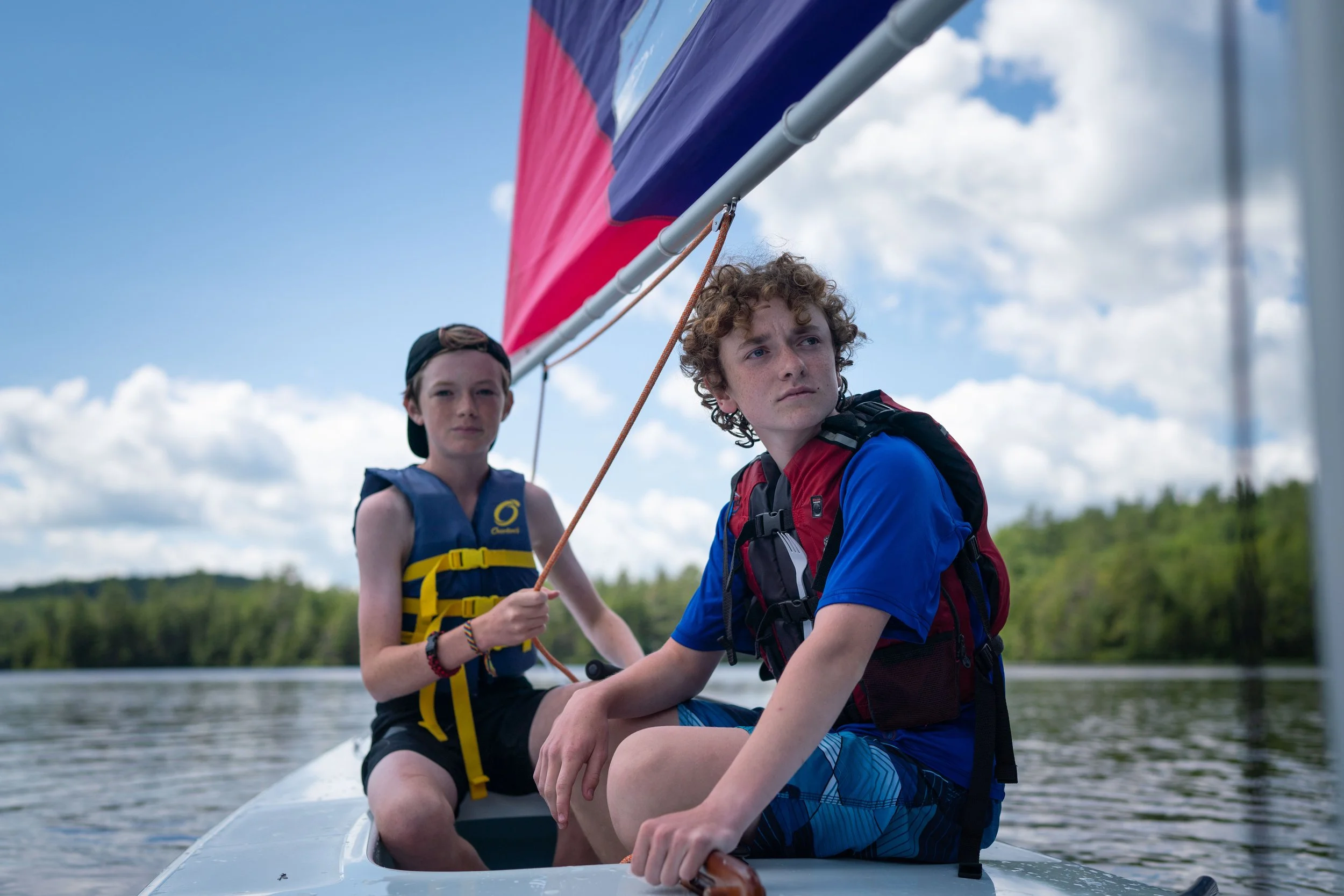 Two young boys sitting on a small sailboat, one holding a sailing rope, with a colorful sail, on a body of water with a tree-lined shore in the background on a partly cloudy day.