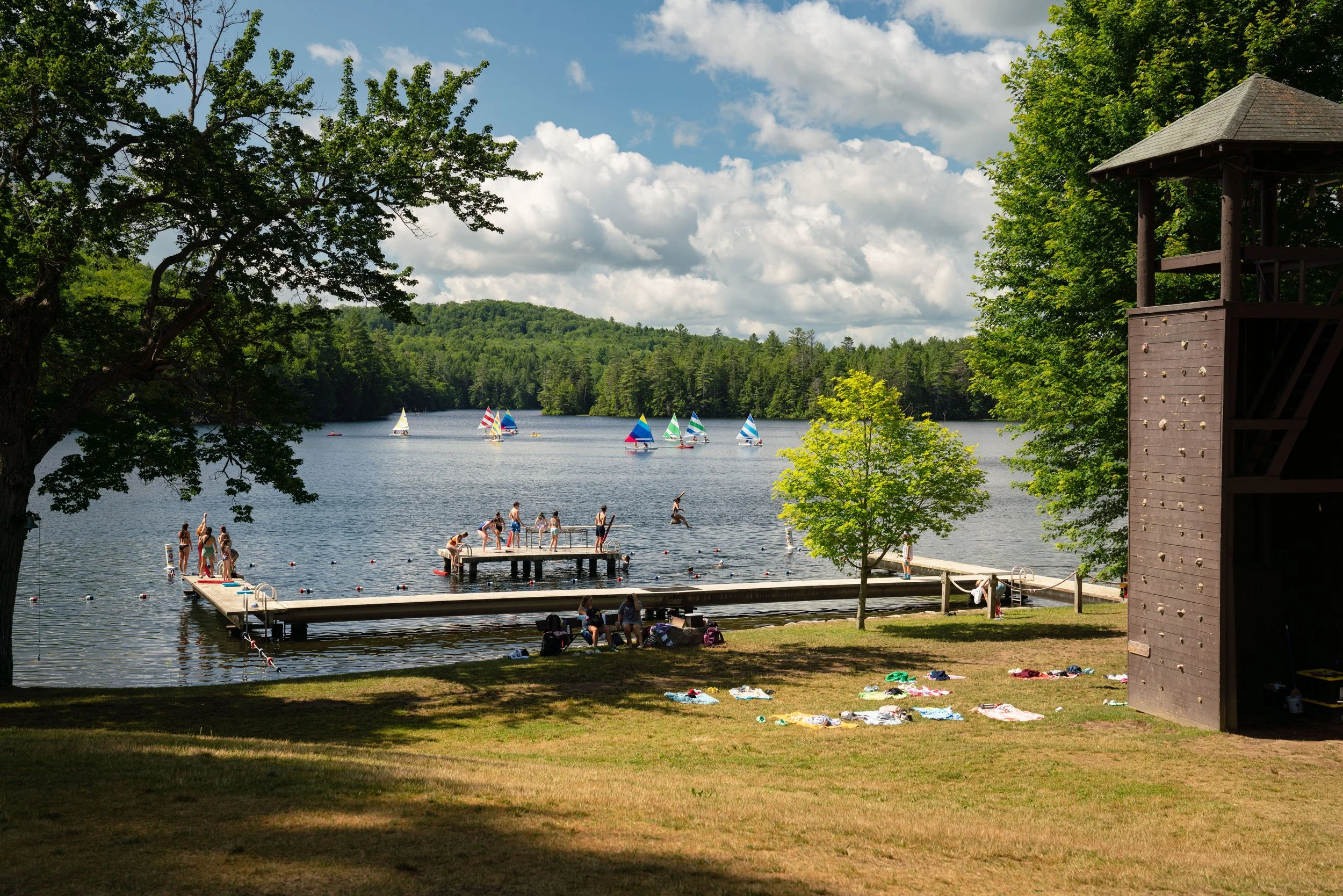 People swimming and playing at a lakeside dock on a sunny day with sailboats on the water and green trees surrounding the lake.