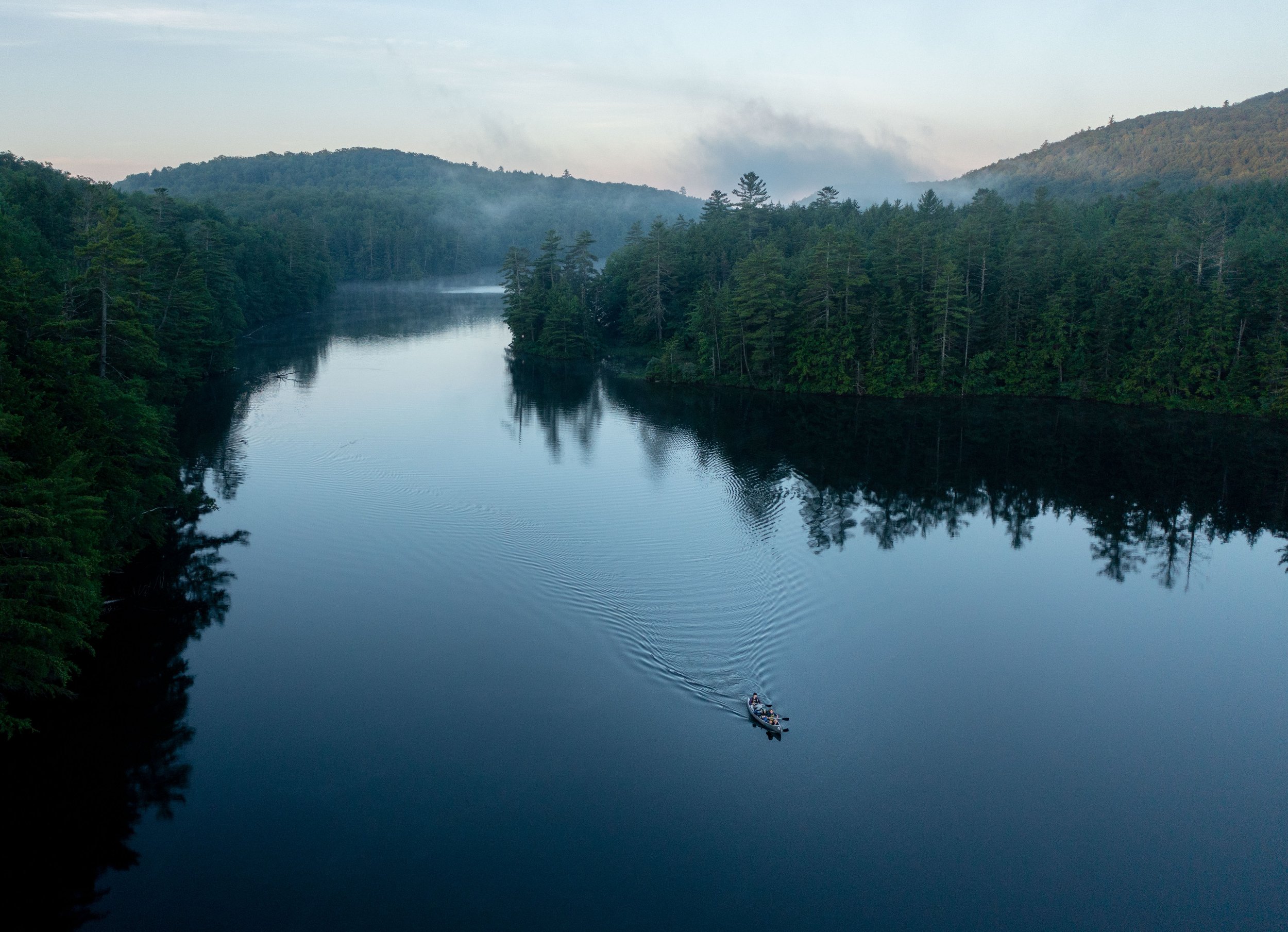 A boat with people on a calm river surrounded by dense green forest and distant hills, with mist or smoke rising in the background.