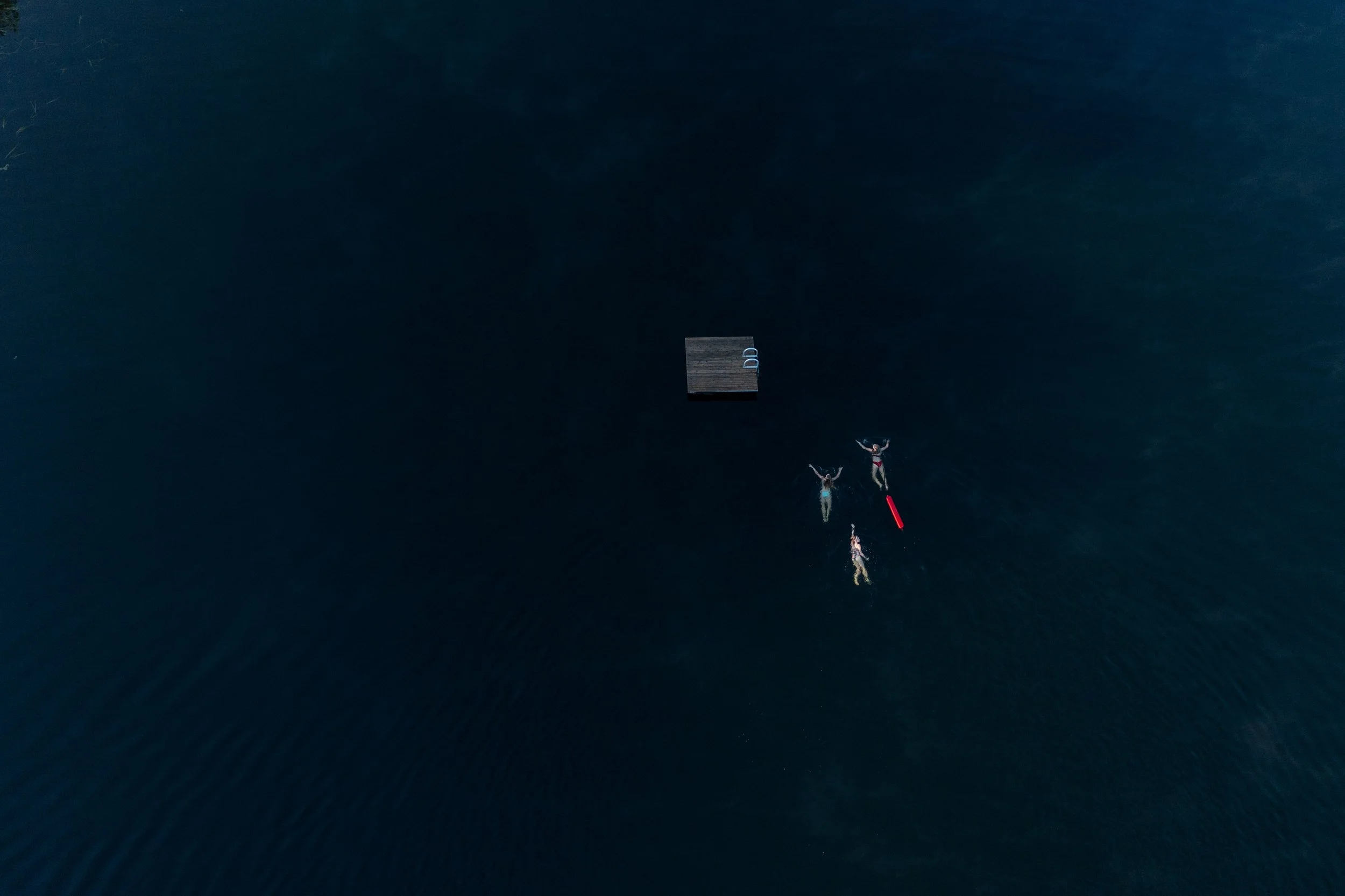 Four people floating on a dark body of water, with a floating platform nearby, aerial view.
