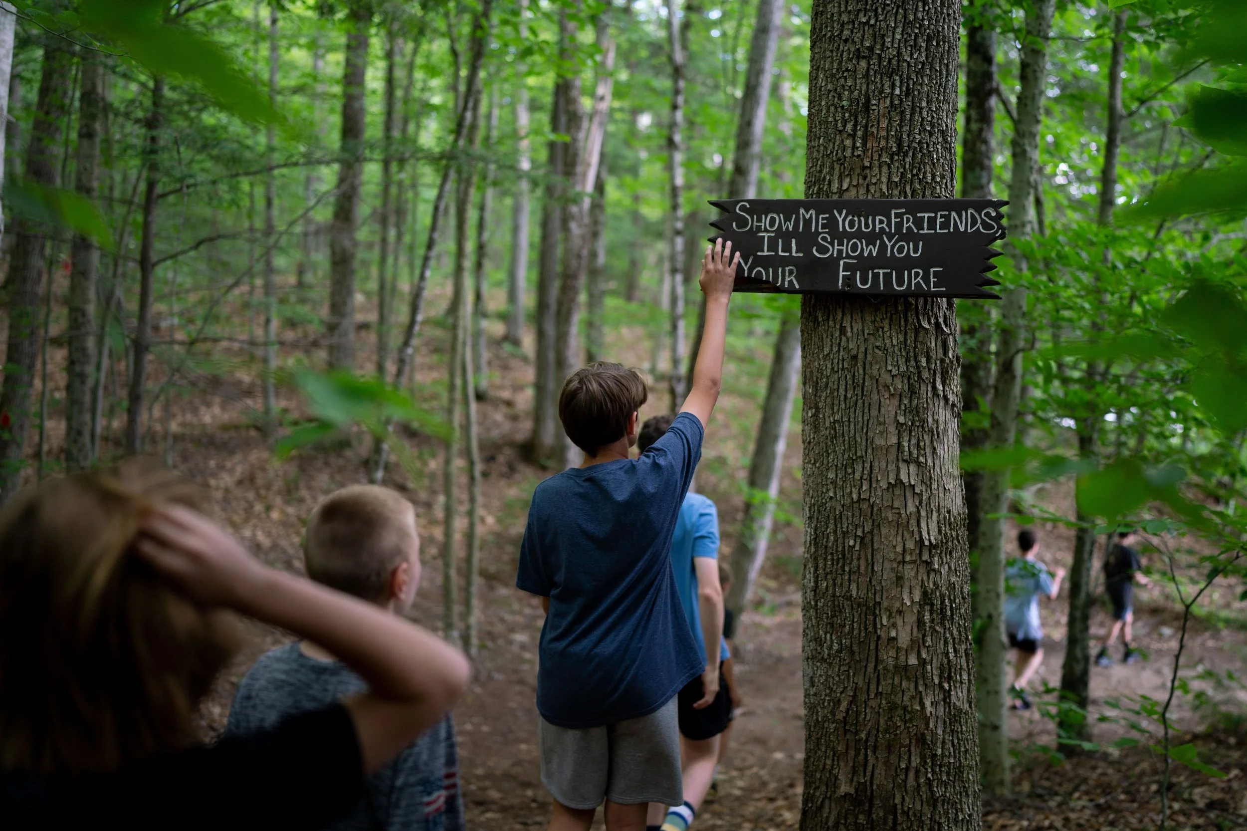 Children walking in a wooded forest, with one boy reaching up to touch a black sign nailed to a tree that says 'Show me your friends, I'll show you your future'.