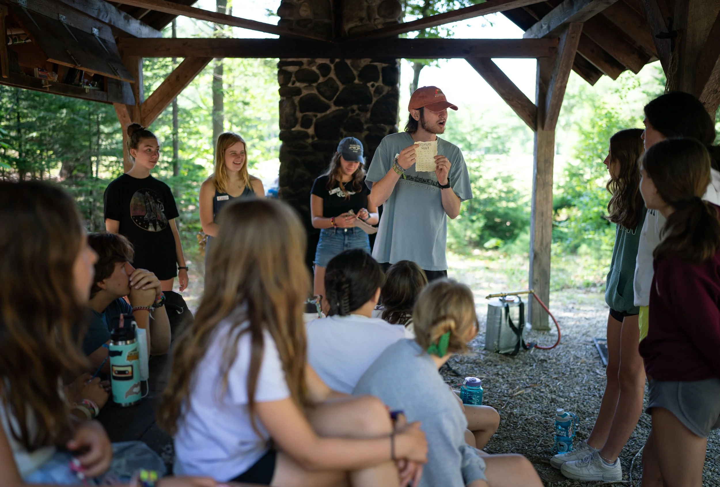 Group of children and teenagers gathered outdoors in a wooden shelter, with a young man in a pink cap holding a piece of paper, appearing to lead or instruct the group during daytime.
