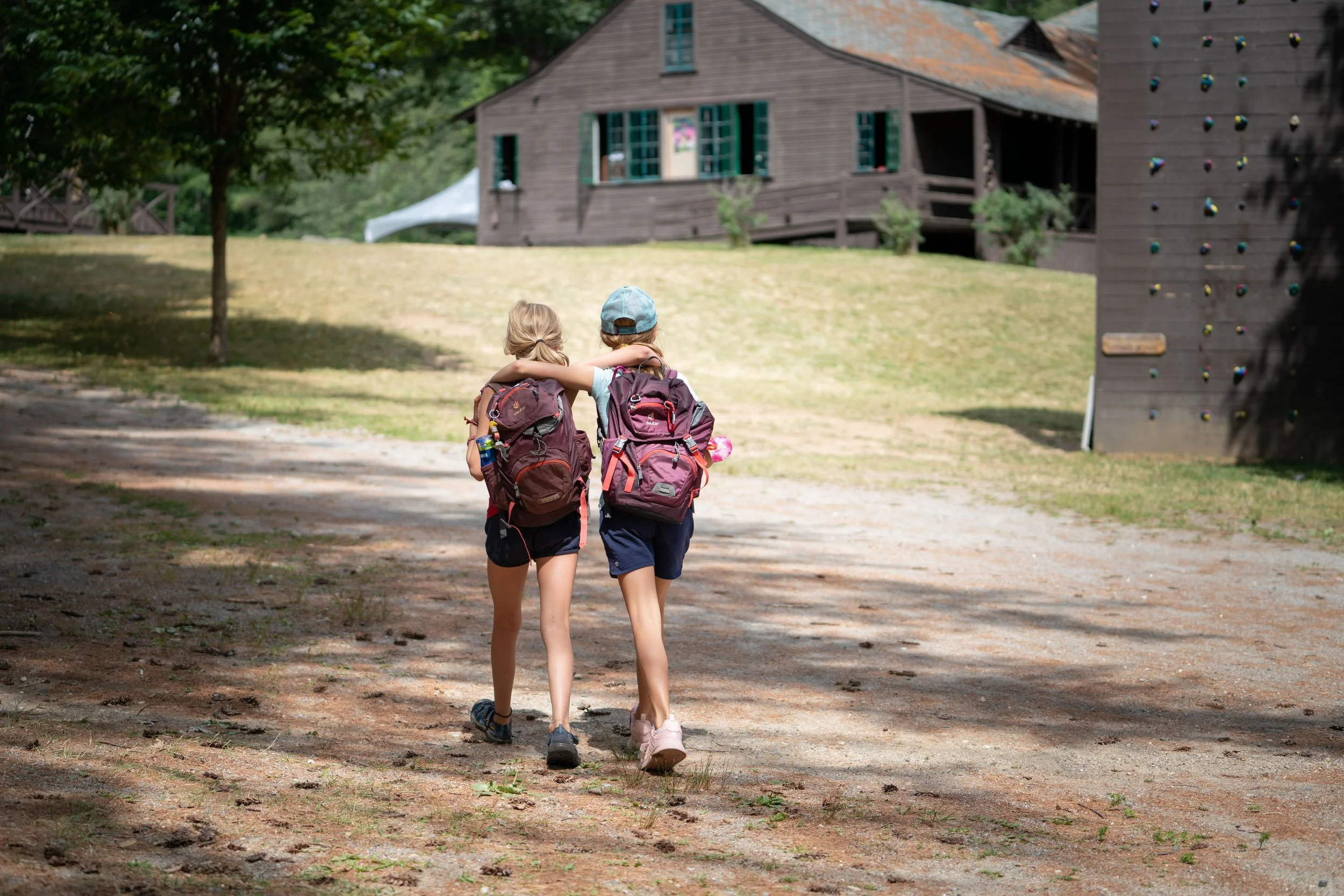 Two children with backpacks walking arm in arm on a dirt path through a park or campground, with a wooden cabin and a climbing wall in the background.