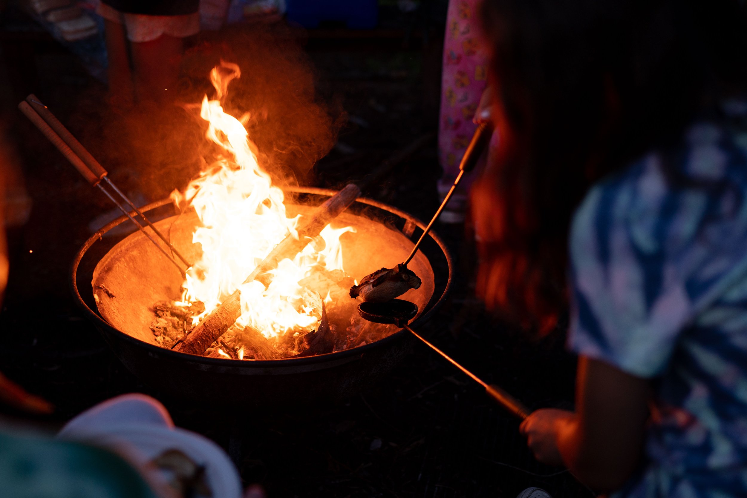Child roasting food over an open fire in a round metal container, using a stick and lying close to the flames at night.