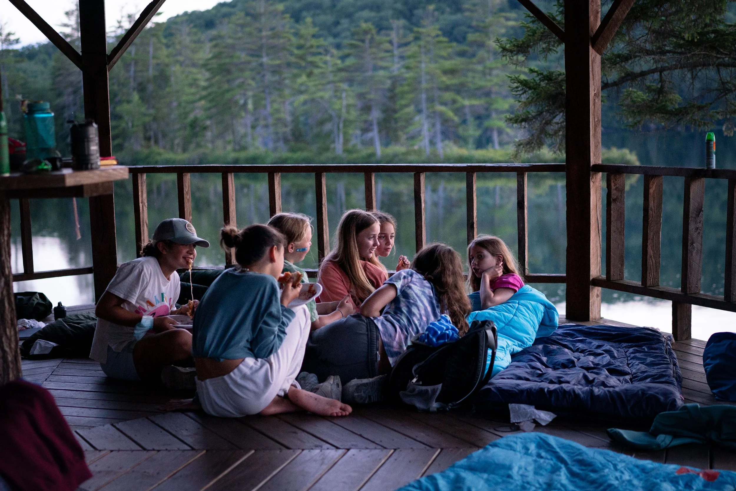Group of children sitting on a wooden deck overlooking a lake surrounded by trees, sharing a snack and talking.