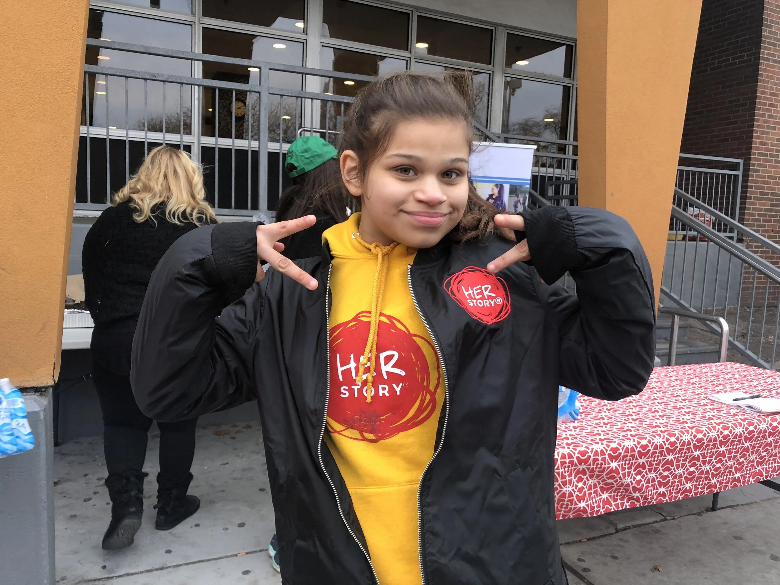 A young girl with brown hair smiling and making a peace sign with both hands at an outdoor event, wearing a yellow shirt and black jacket, with a table covered in red patterned tablecloth in front of her. There are other people and a building with large windows in the background.