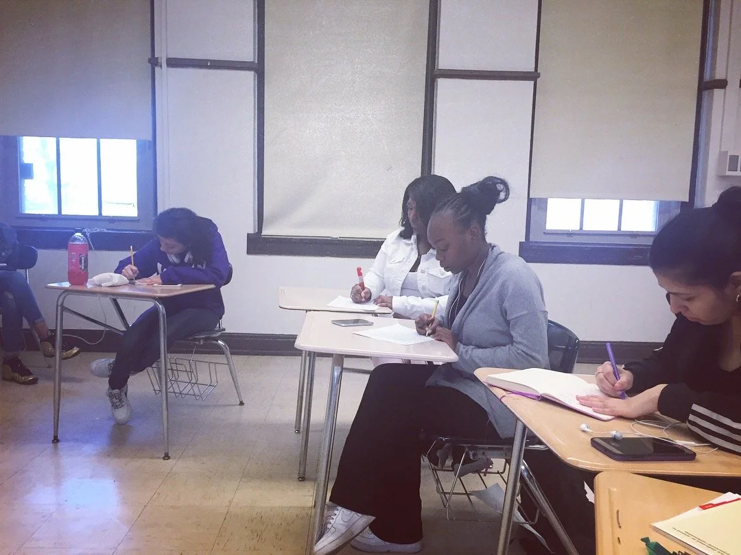 Four students sitting at desks in a classroom, writing in notebooks, with windows and beige walls in the background.